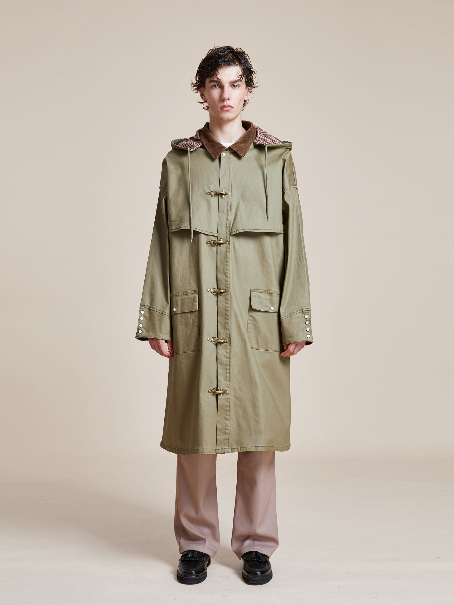 A person standing and facing forward, wearing a long, olive green Prairie Buckle Coat by Found with a hood, reminiscent of traditional 80's hunting jackets, beige pants, and black shoes. The background is plain and neutral.