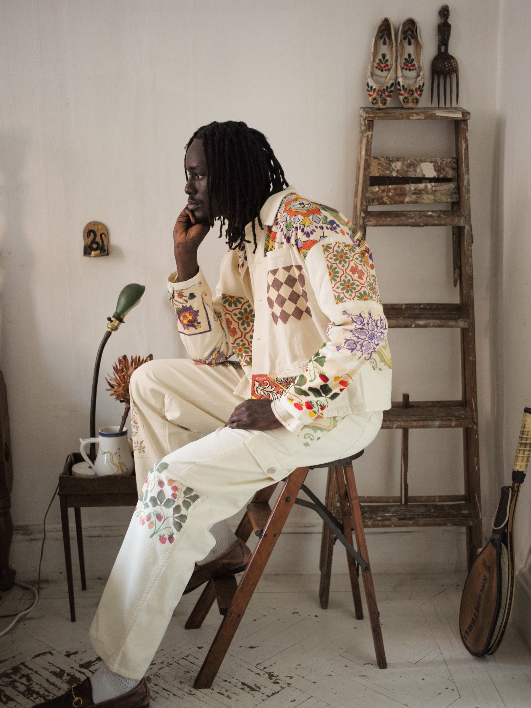 A person is seated on a stool, clad in a white Mosaic Work Jacket from FOUND, featuring colorful floral patterns with Indo-Aryan flair. A ladder, lamp, and decorative items offer an artful backdrop.