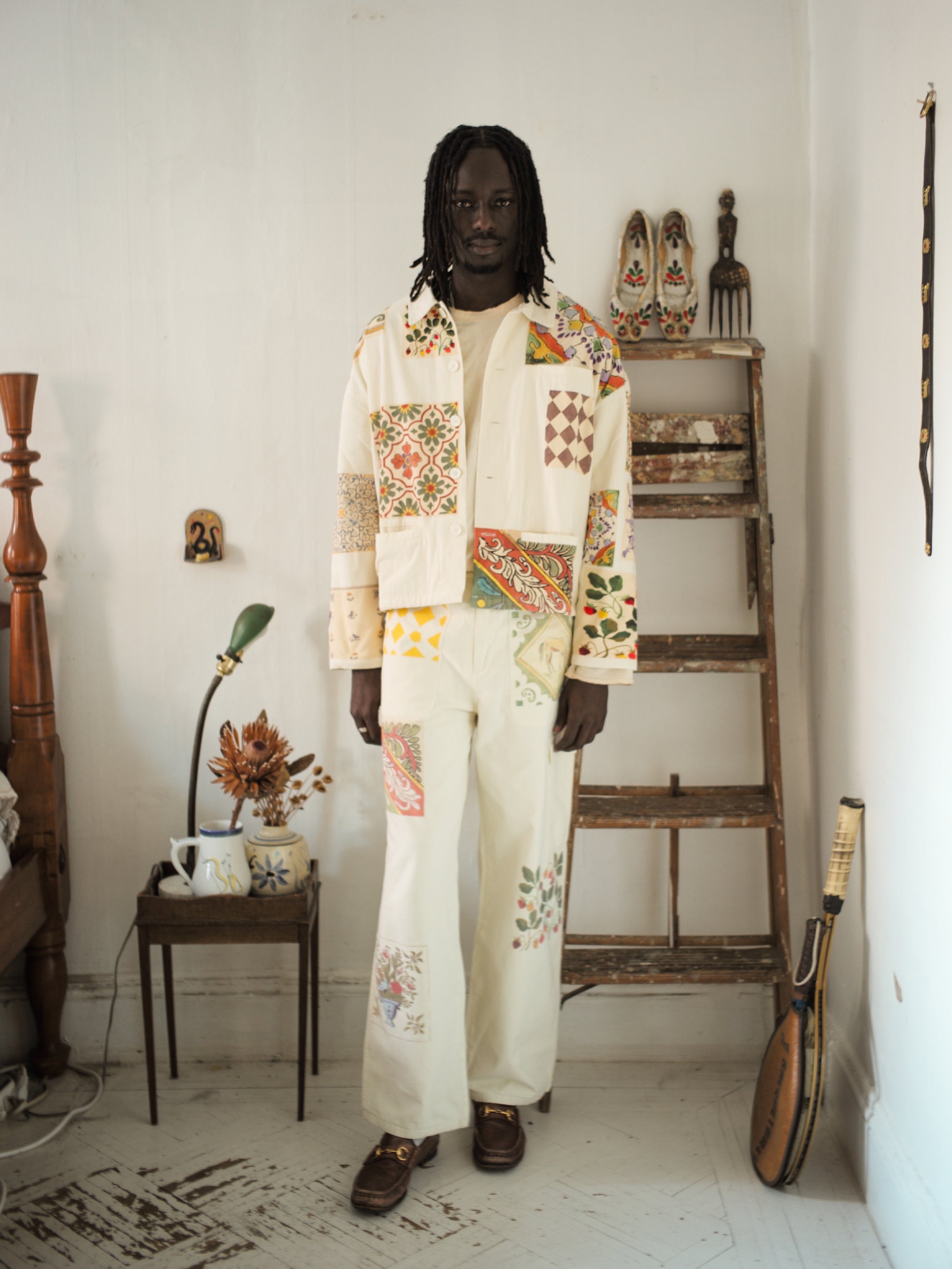 A person stands in a room dressed in an off-white outfit with a FOUND Mosaic Work Jacket, beside a wooden ladder with decor, and a table displaying a vase and mug.