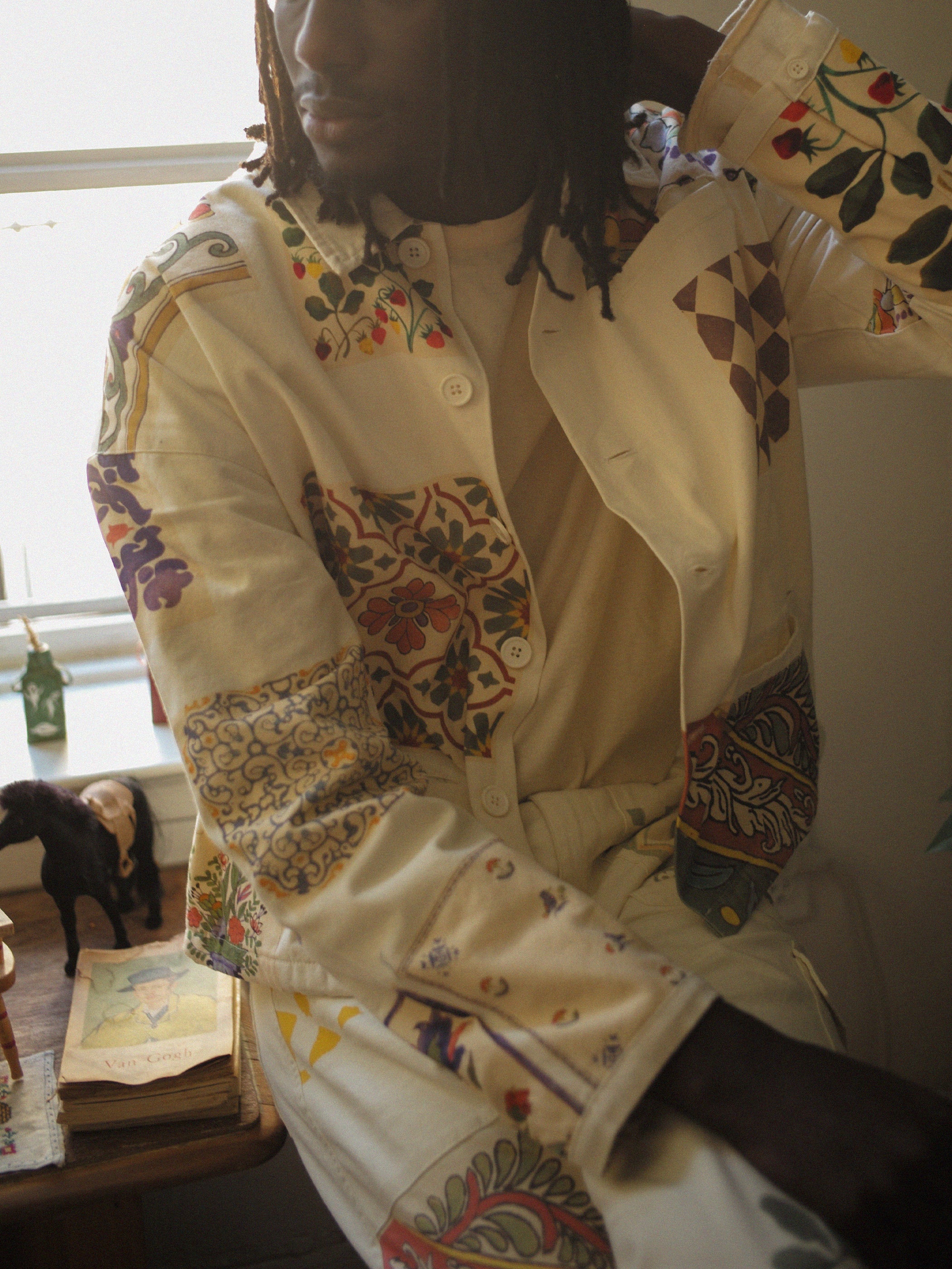 A person in a room wearing the FOUND Mosaic Work Jacket, featuring colorful Indo-Aryan patterns, sits on a table beside a small statue and books.