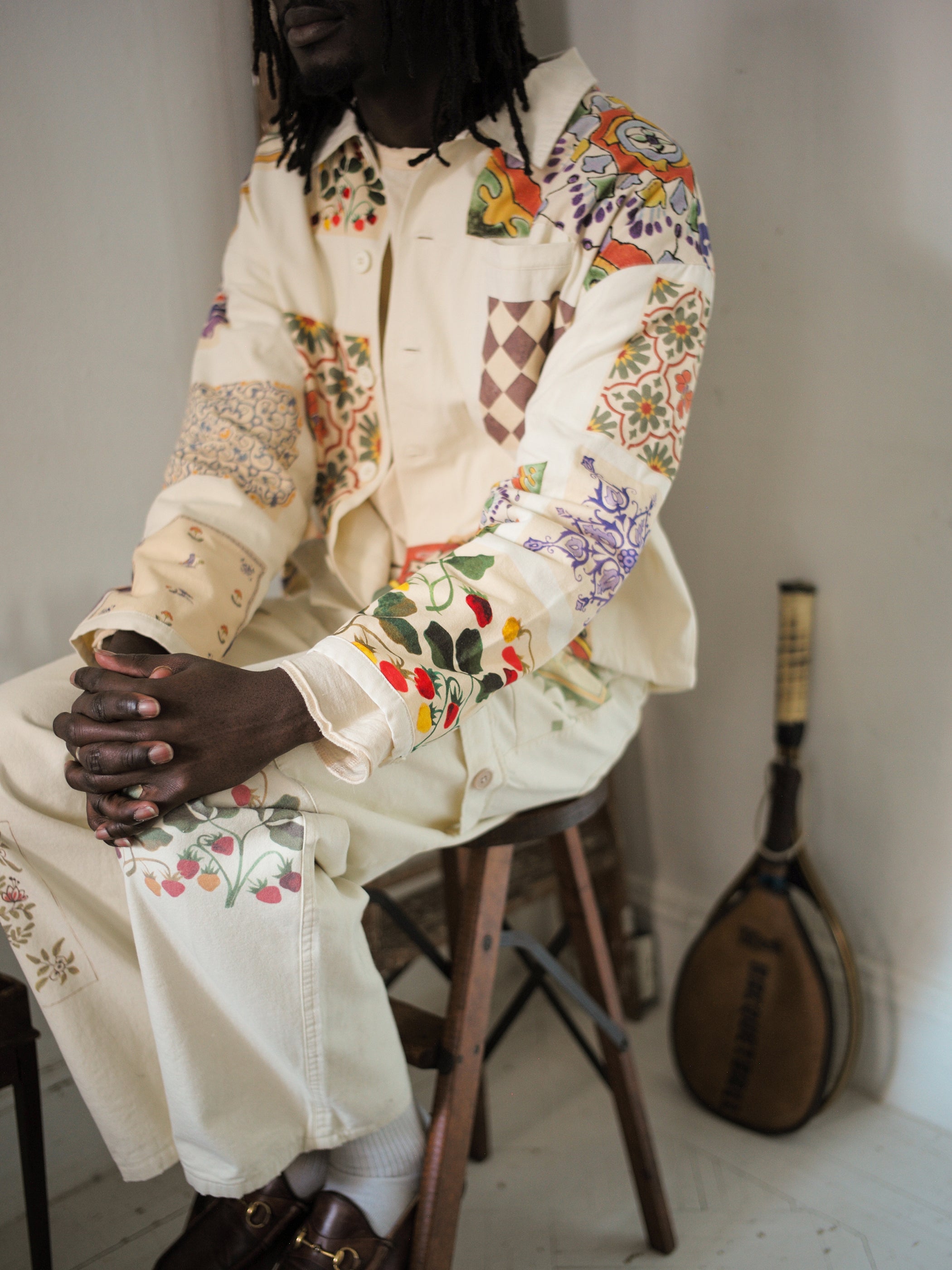 Wearing Profound's Mosaic Pants, a person sits on a stool, dressed in cream attire with vibrant Indo-Aryan-inspired patches. A musical instrument casually leans against the wall in the background.