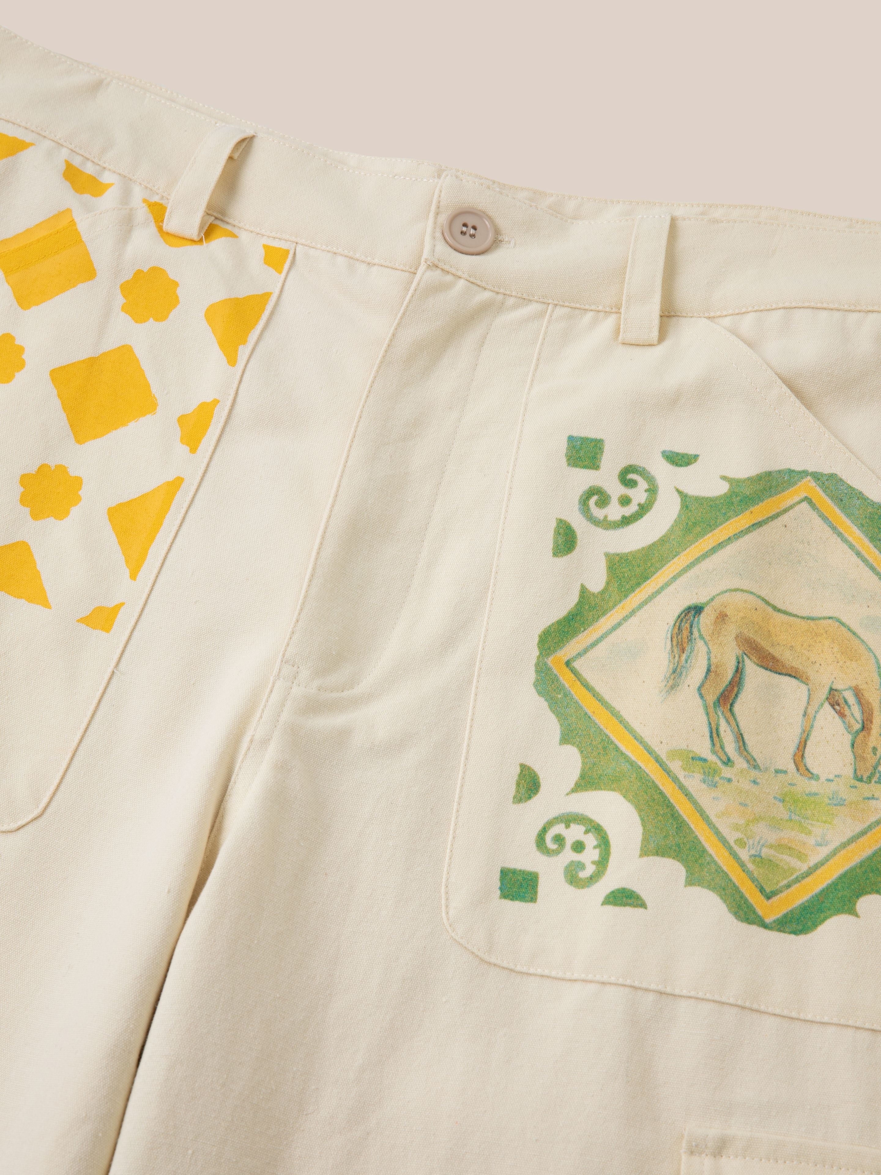 A close-up of cream-colored Mosaic Pants by Profound featuring a button closure and two printed designs: yellow geometric shapes on one side and a green-bordered image of a horse on the other, all showcased on durable cotton canvas fabric.