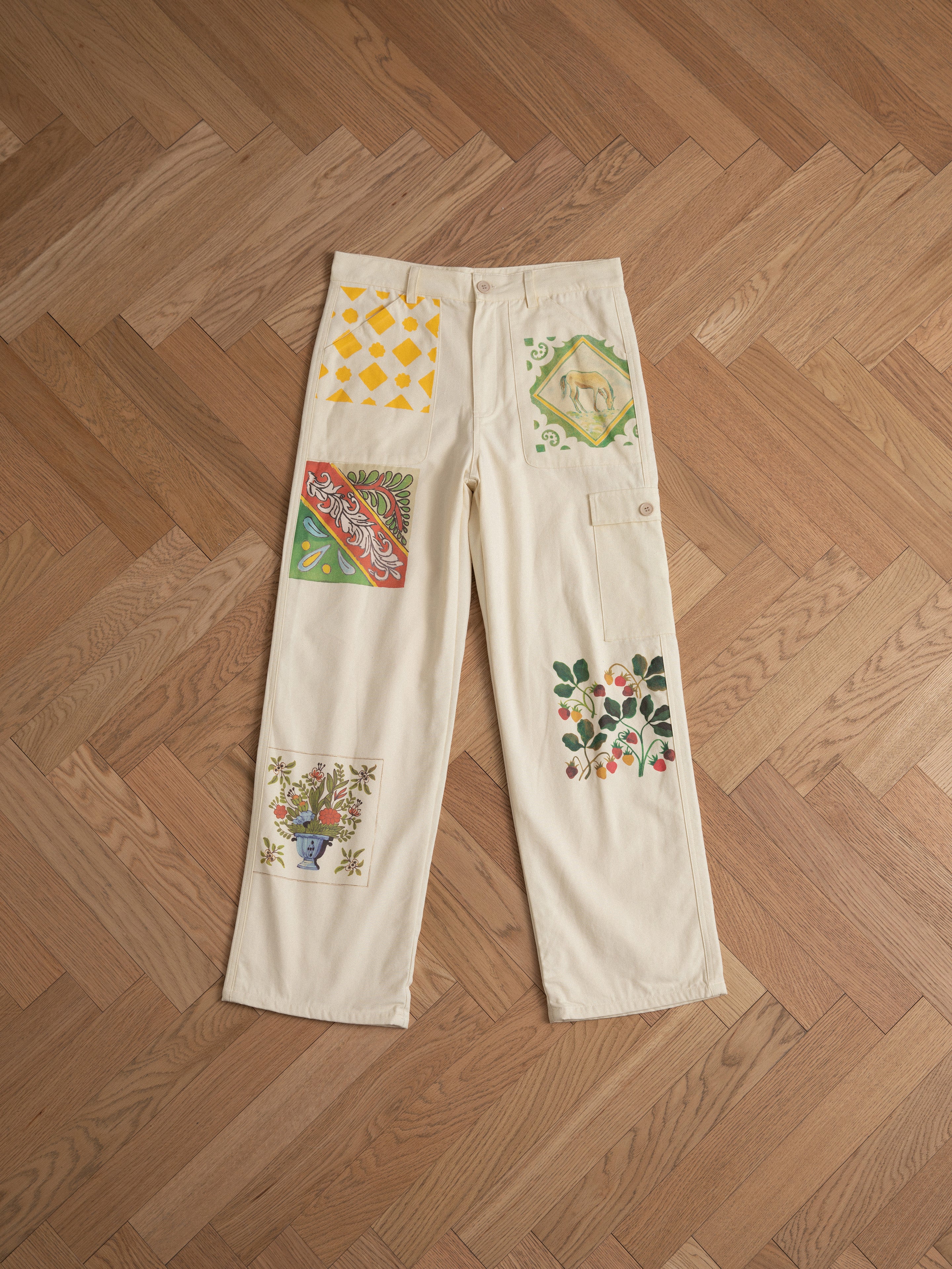 Mosaic Pants by Profound, with various colorful designs and patches reminiscent of Indo-Aryan patterns, laid flat on a wooden herringbone-patterned floor.