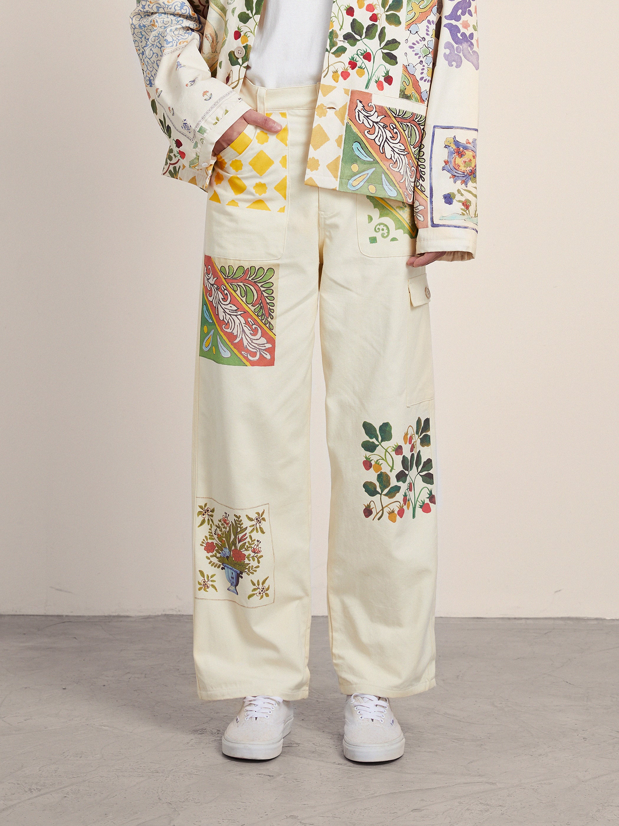 Person wearing a colorful patchwork jacket and **Profound Mosaic Pants** with floral and geometric designs reminiscent of Indo-Aryan patterns, standing on a concrete floor. White sneakers are partially visible.