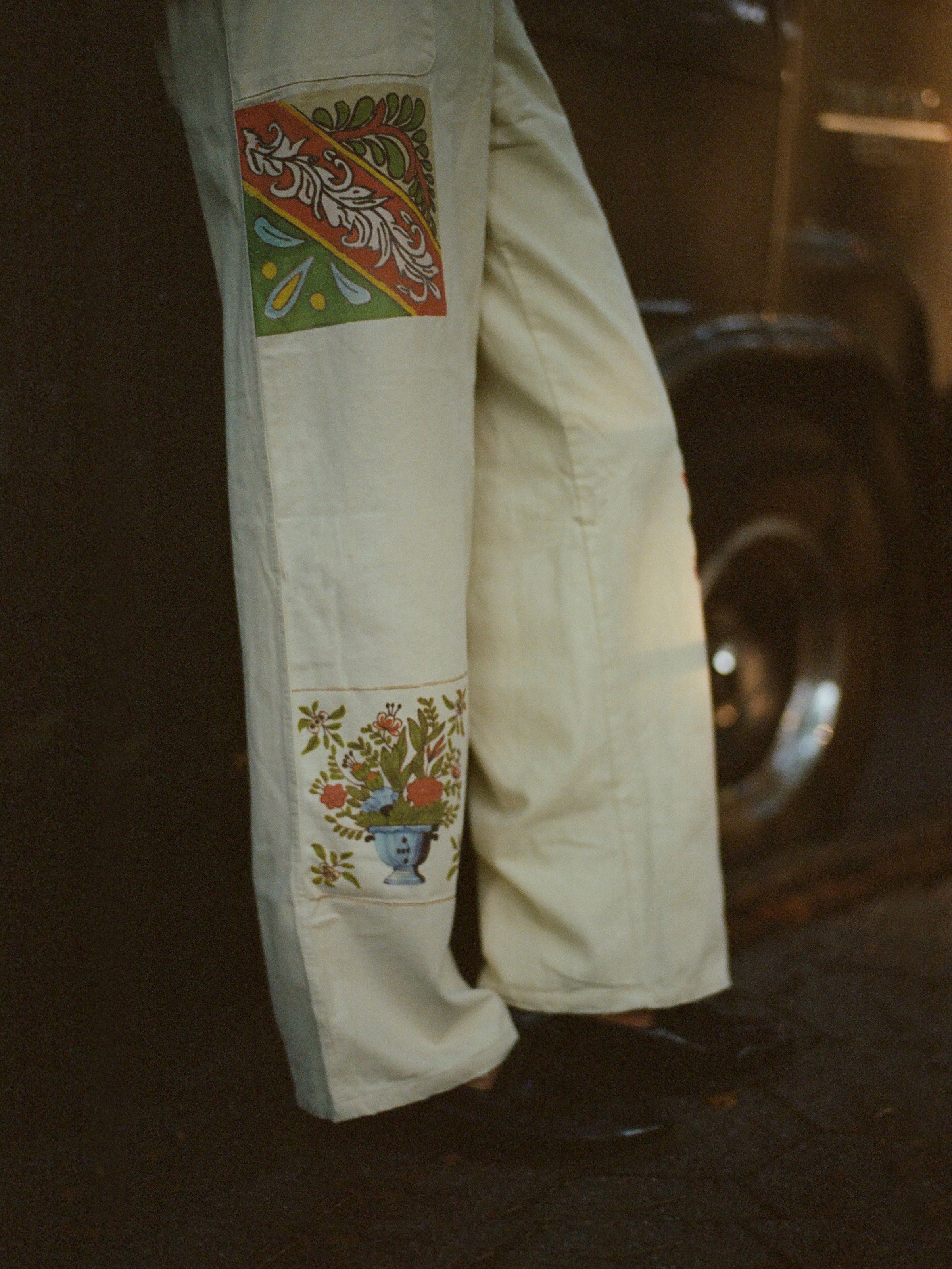 A person wearing cream-colored Profound Mosaic Pants with colorful rectangular embroidered patches of a floral design, reminiscent of traditional Indo-Aryan patterns, stands beside a vehicle tire.