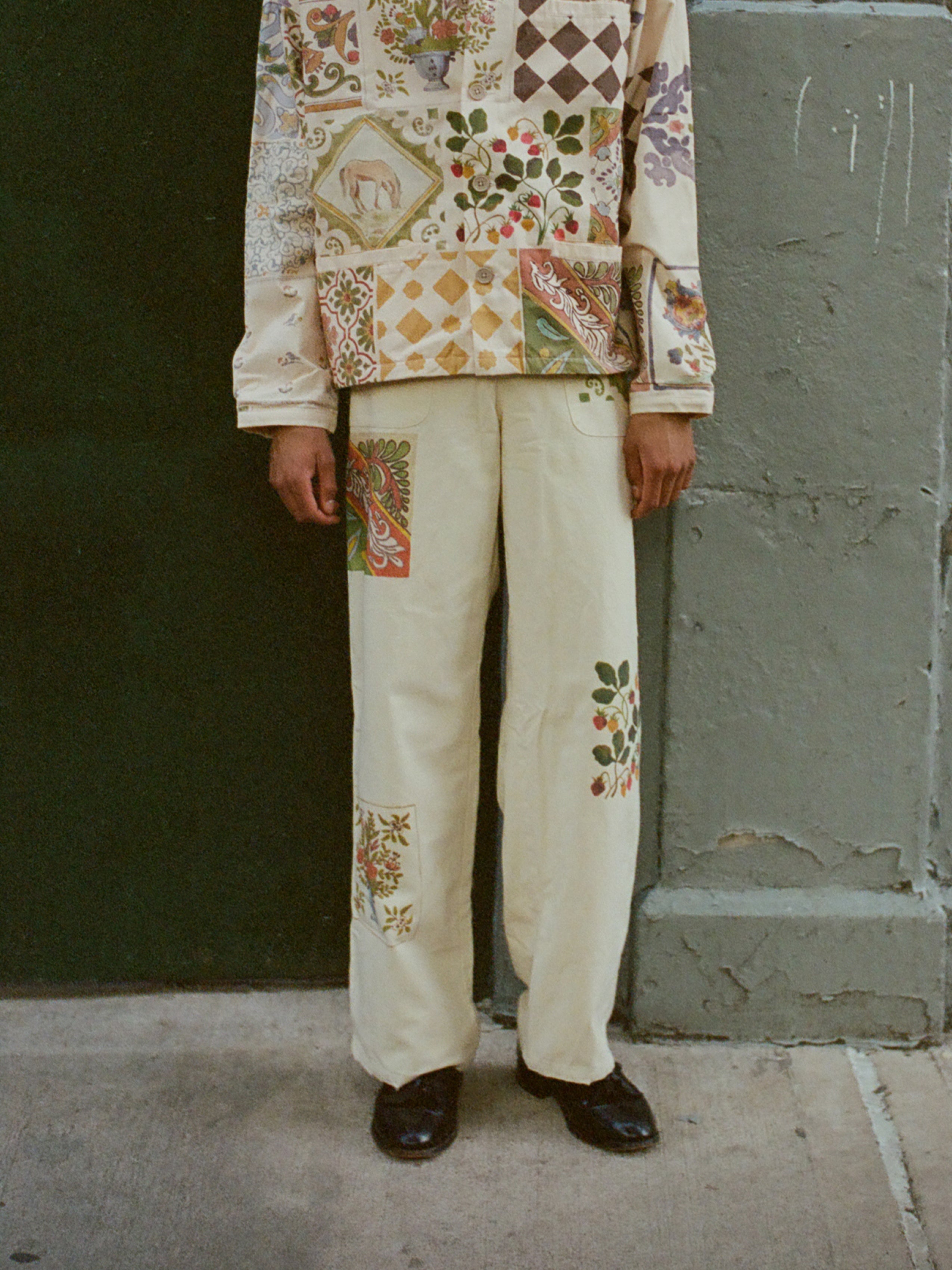 A person stands against a green and grey wall, wearing a cream-colored outfit with various colorful floral and geometric patch designs. The vibrant Mosaic Pants from Profound, made from cotton canvas fabric, feature intricate traditional Indo-Aryan patterns. Only the lower half of the person's body is visible.