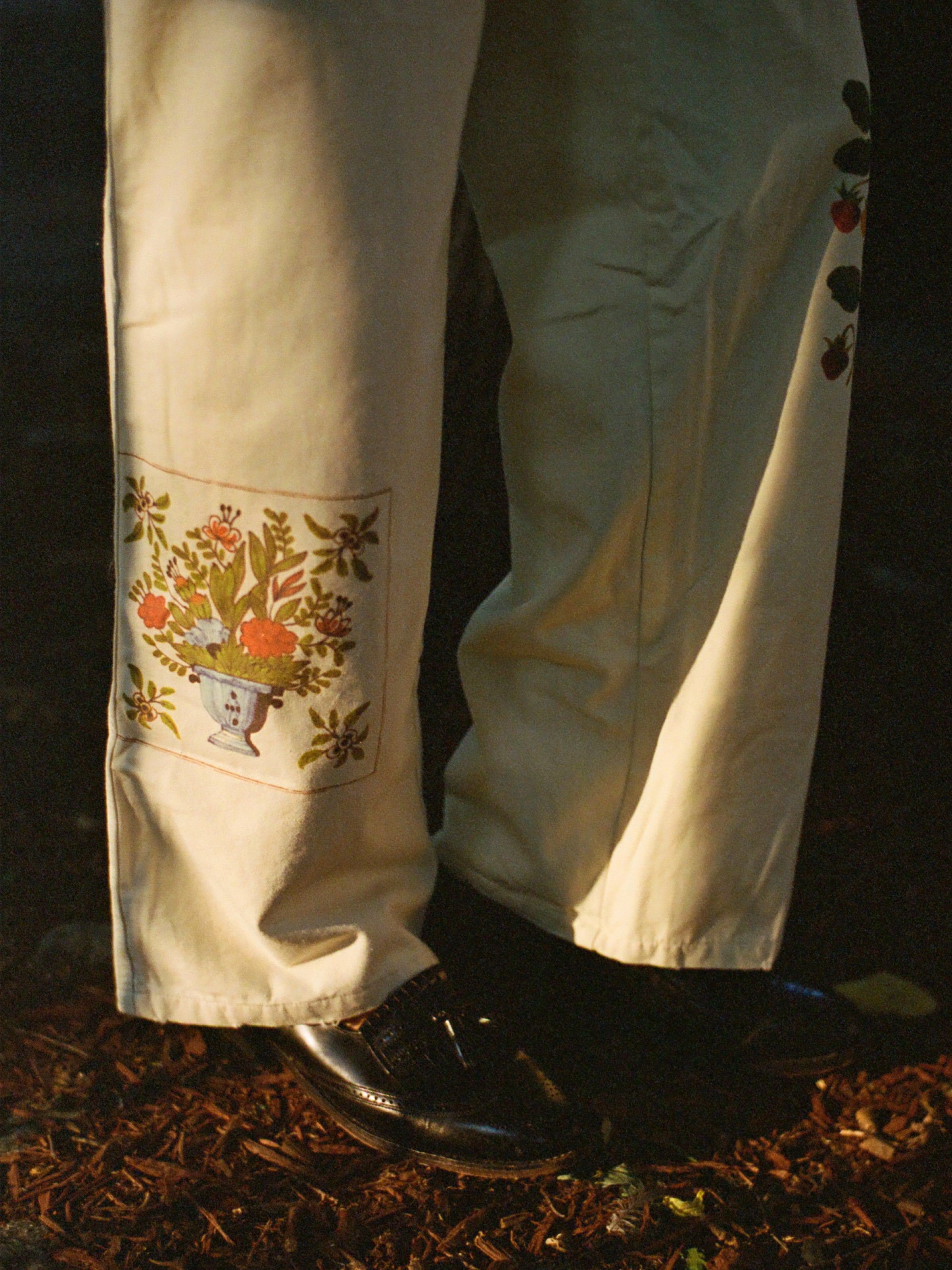 Close-up of a person wearing Profound Mosaic Pants crafted from breathable cotton canvas, featuring traditional Indo-Aryan patterns near the bottom and black shoes, standing on a dark brown, bark-covered ground.