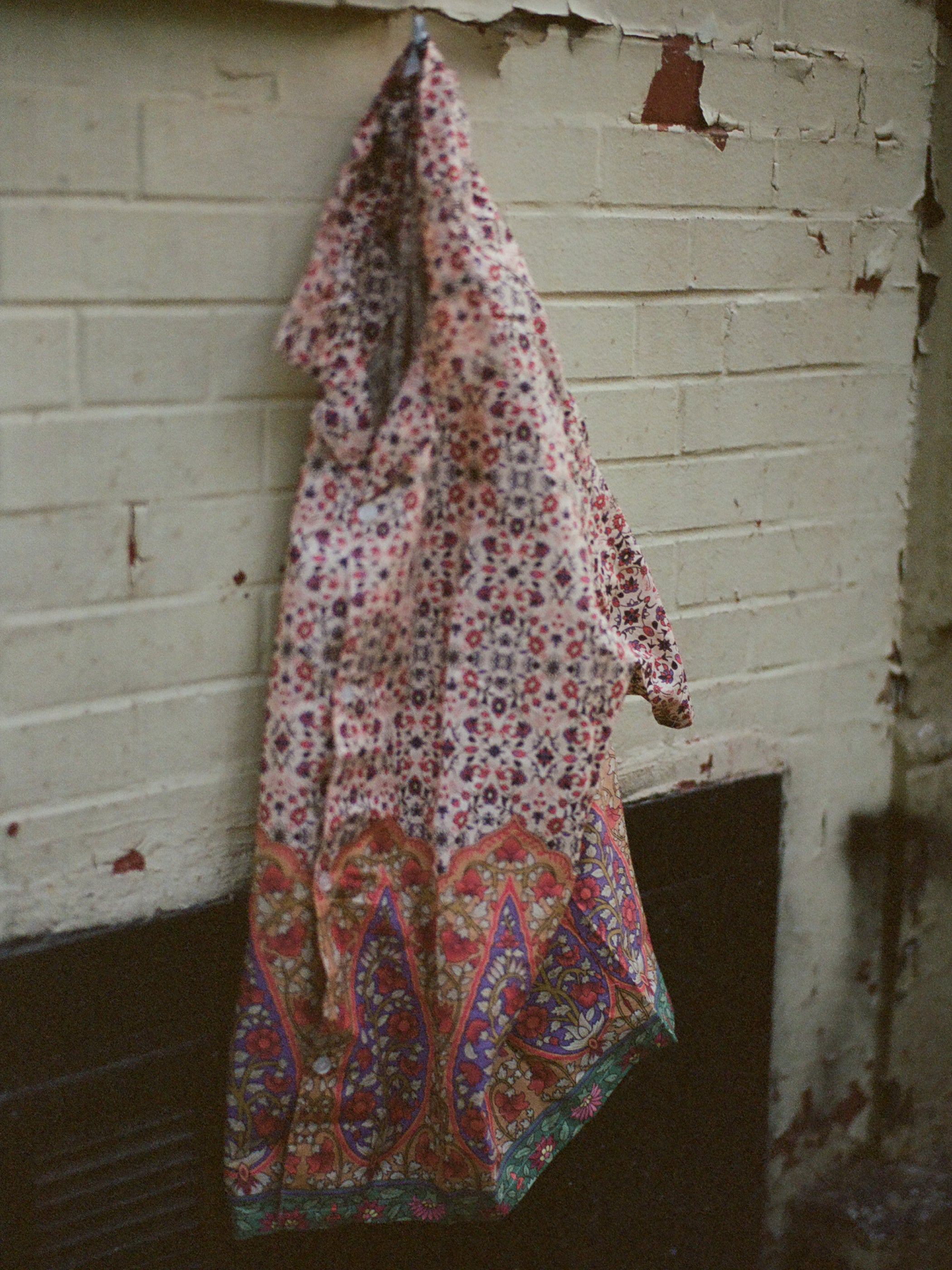 A colorful, patterned piece of 100% cotton fabric hangs on a hook against a faded, chipped brick wall, reminiscent of traditional Indo patterns seen in a Mughal Camp Shirt by Profound.