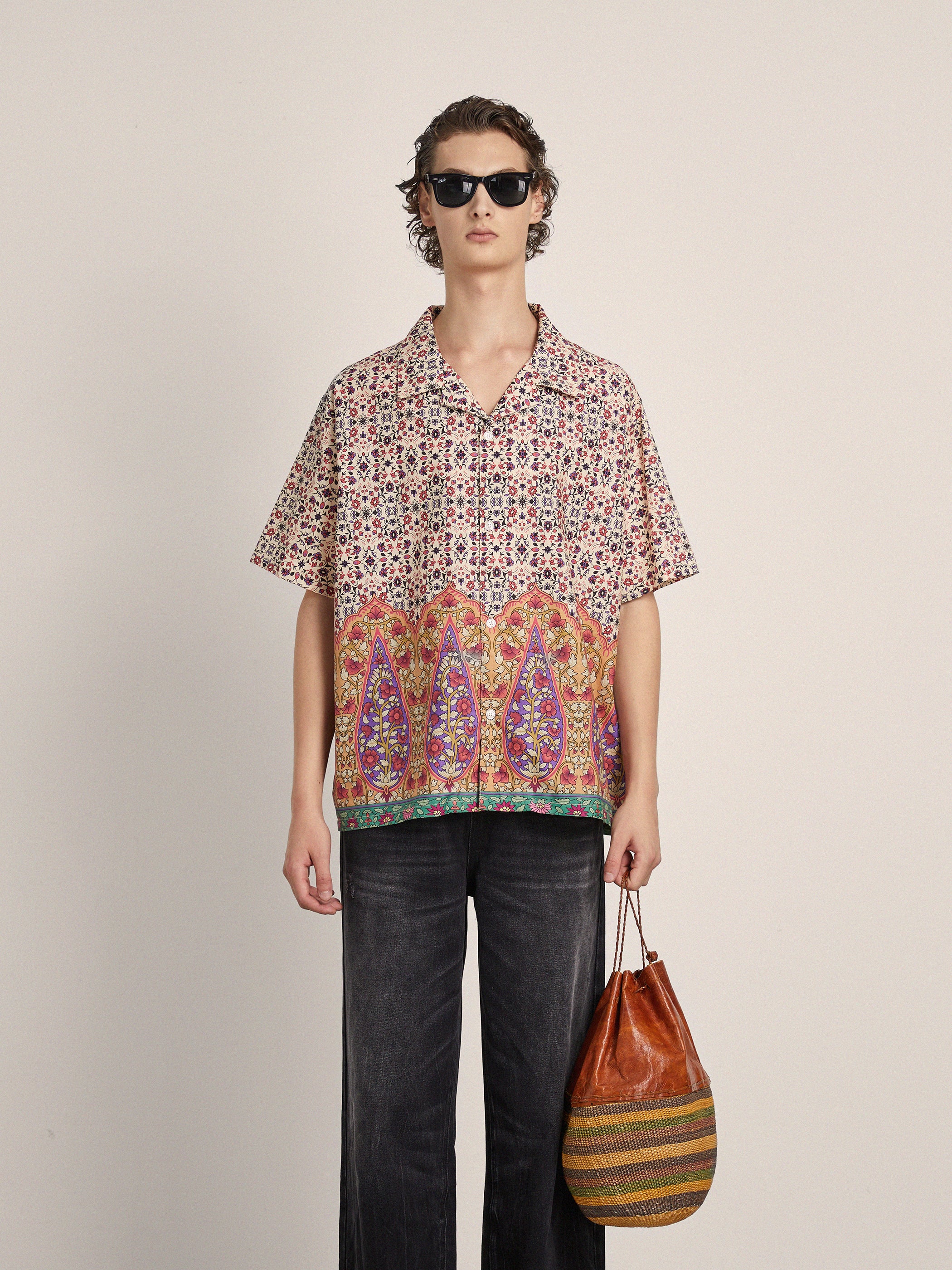 A person wearing sunglasses, a Profound Mughal Camp Shirt with intricate Indo patterns, and dark pants holds a multicolored bag against a plain background.