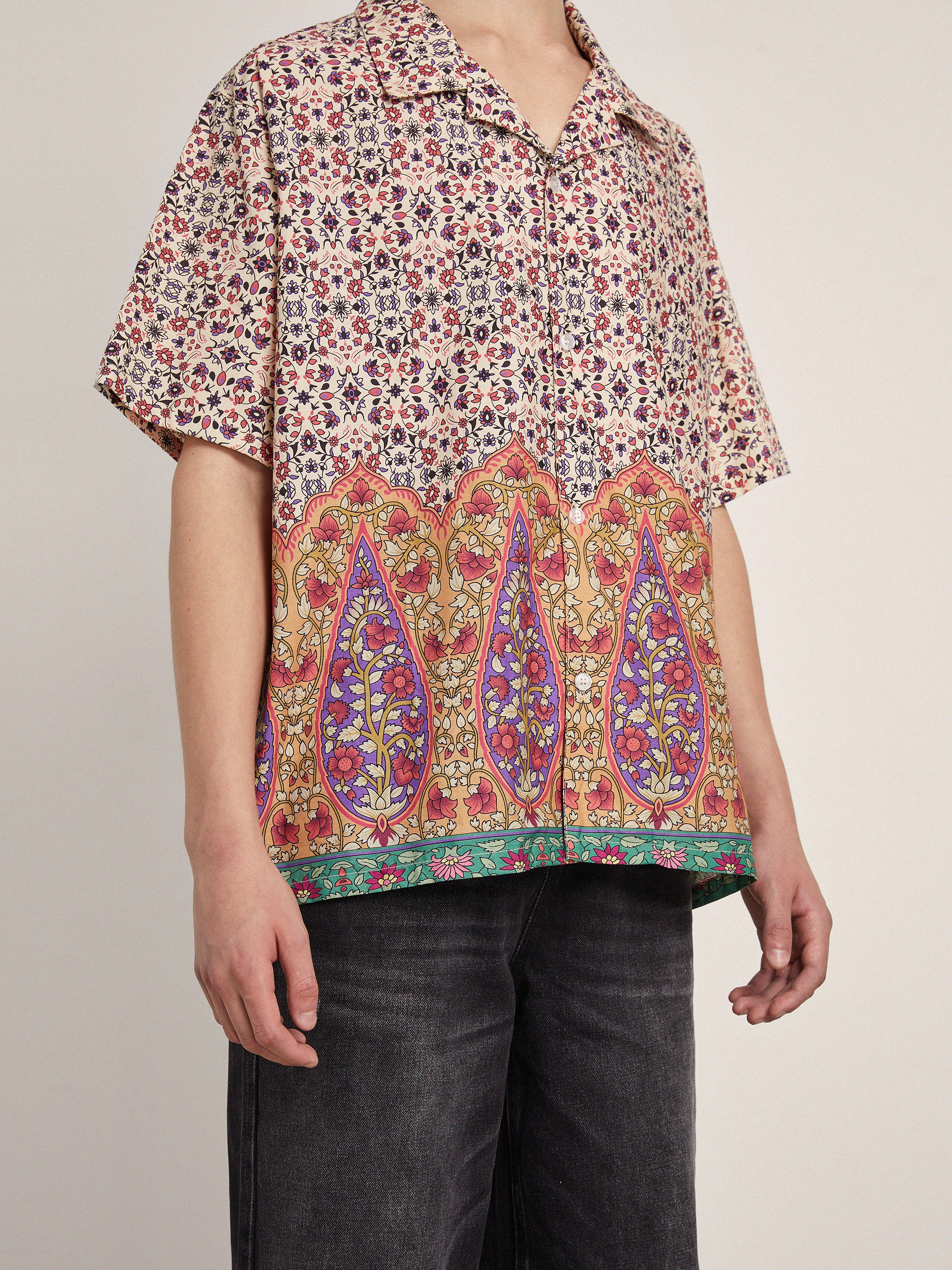 A person is wearing a colorful, patterned short-sleeved Mughal Camp Shirt by Profound with dark jeans. The shirt, made of 100% cotton fabric, features intricate floral and geometric designs inspired by traditional Indo patterns.