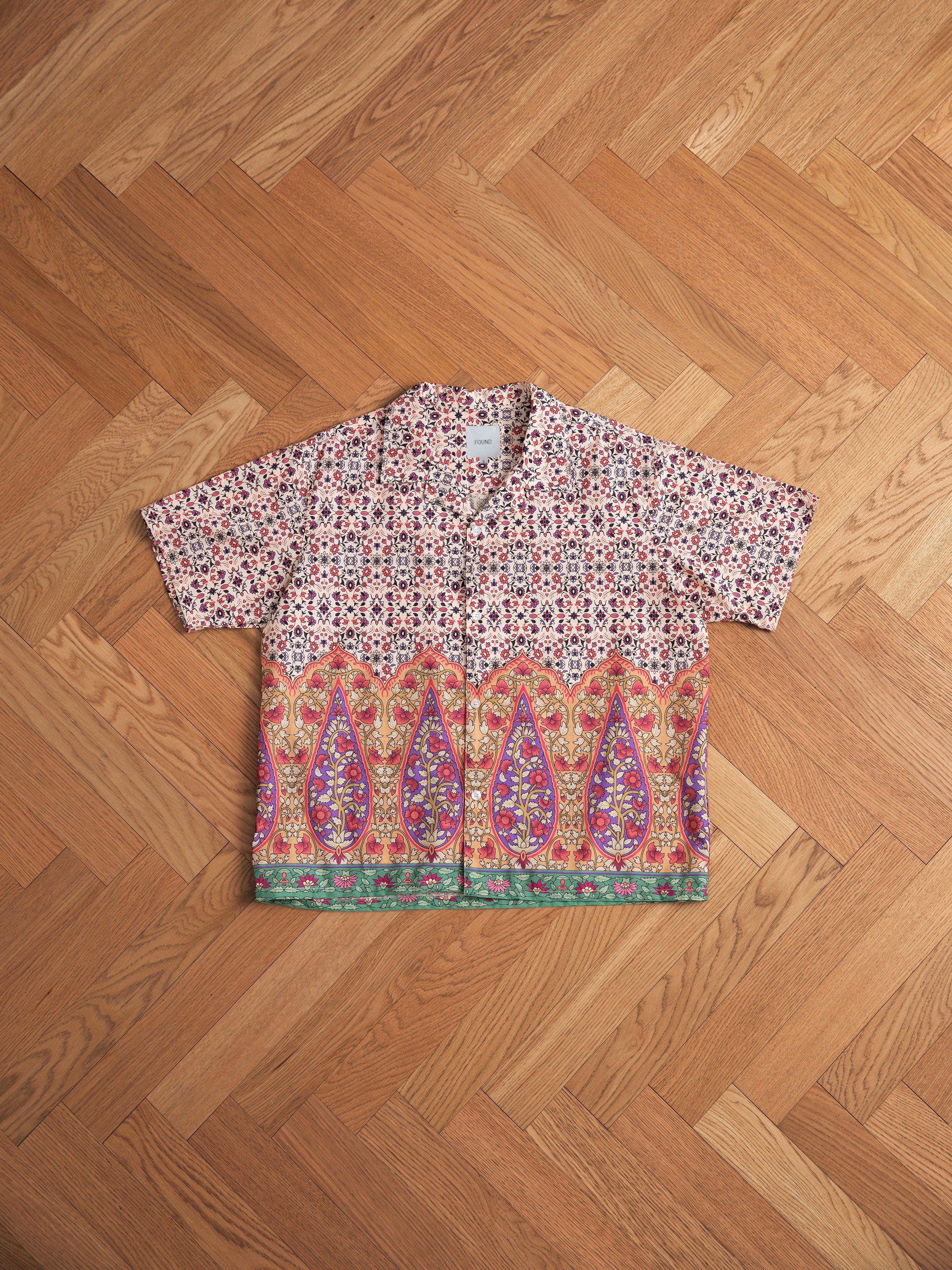 A Mughal Camp Shirt featuring traditional Indo patterns, this short-sleeve piece boasts a vibrant multicolored paisley and geometric design, crafted from 100% cotton fabric and laid flat on a wooden parquet floor. The shirt is by Profound.