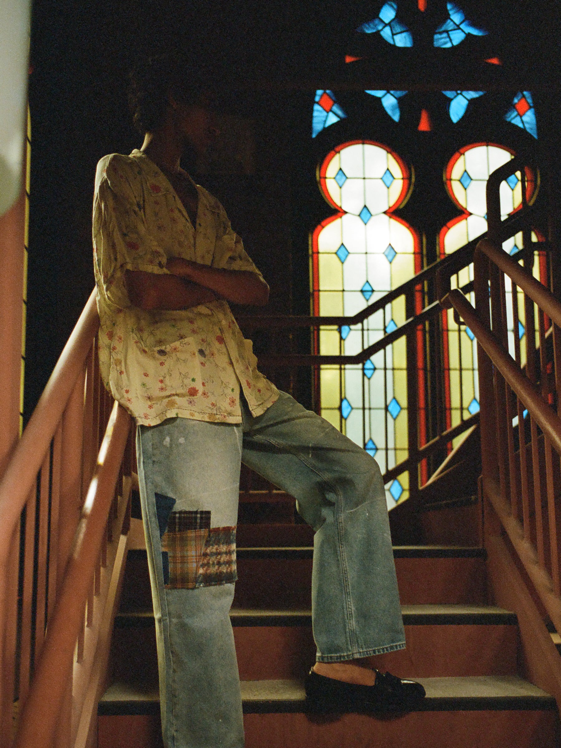 A person stands with arms crossed on a staircase inside a building, wearing a lightweight rayon Multi Floral Camp Shirt by Profound and patched jeans. A stained glass window is visible in the background.