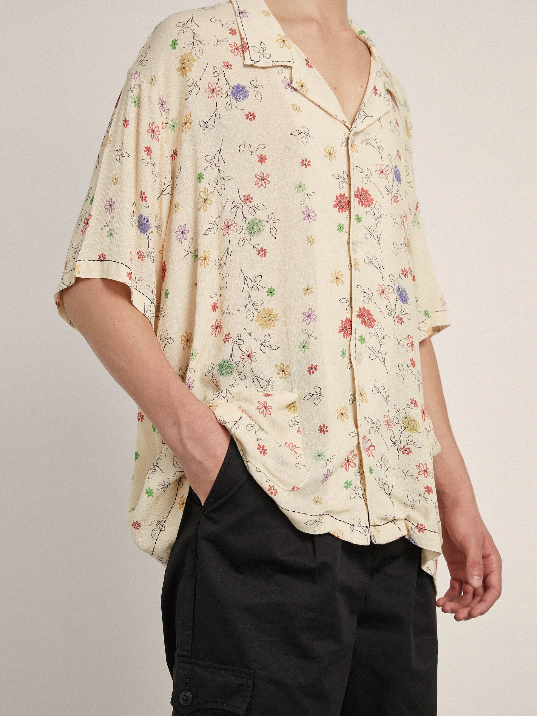 Person wearing a cream-colored, short-sleeved Multi Floral Camp Shirt by Profound paired with black pants. Their left hand is in the pants pocket.