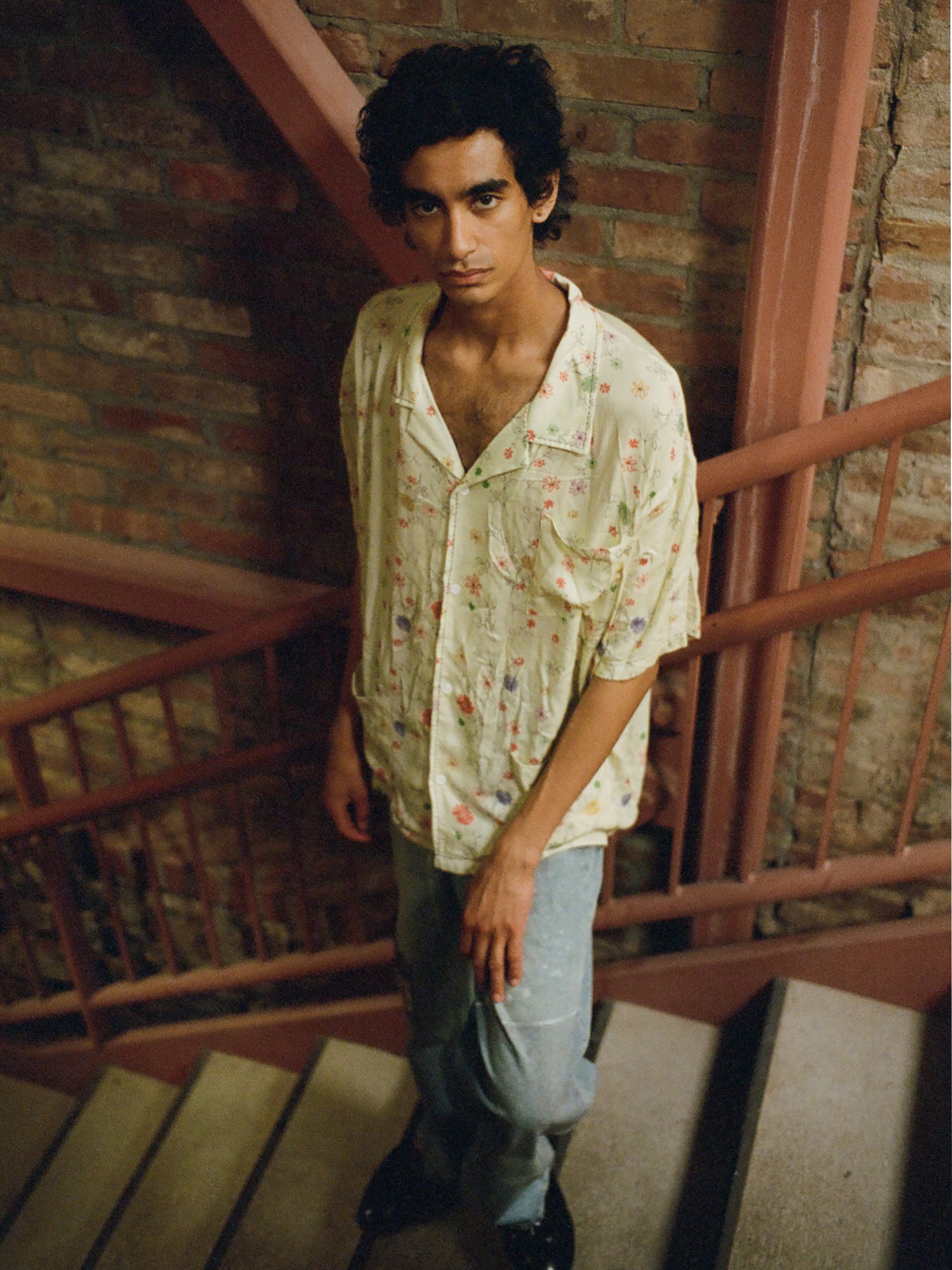 A person with dark curly hair and a neutral expression stands on a staircase against a brick wall, wearing a lightweight rayon Multi Floral Camp Shirt by Profound and blue jeans.