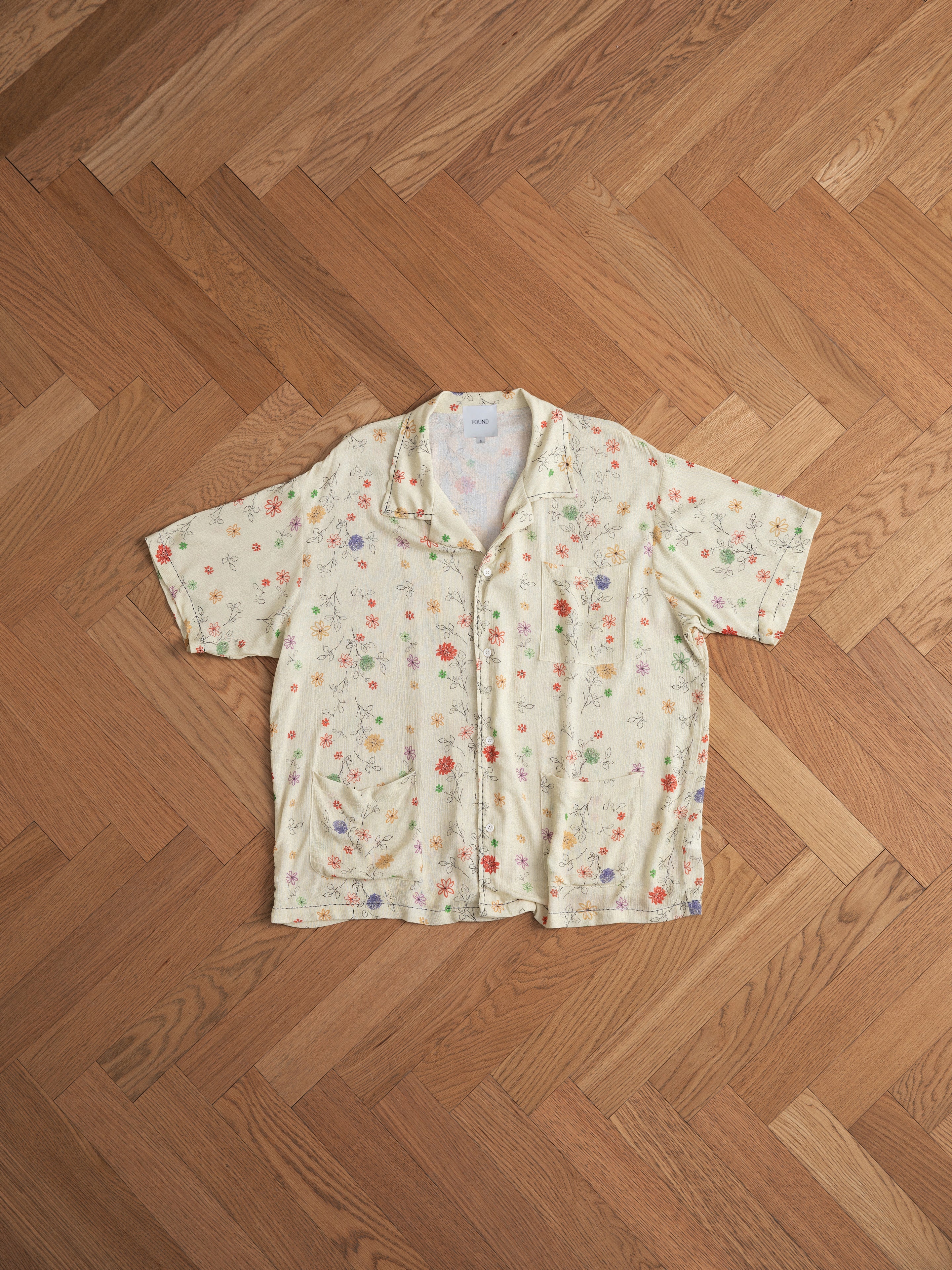 A lightweight rayon Multi Floral Camp Shirt by Profound is laid flat on a wooden herringbone floor, showcasing its short sleeves and vibrant floral pattern.