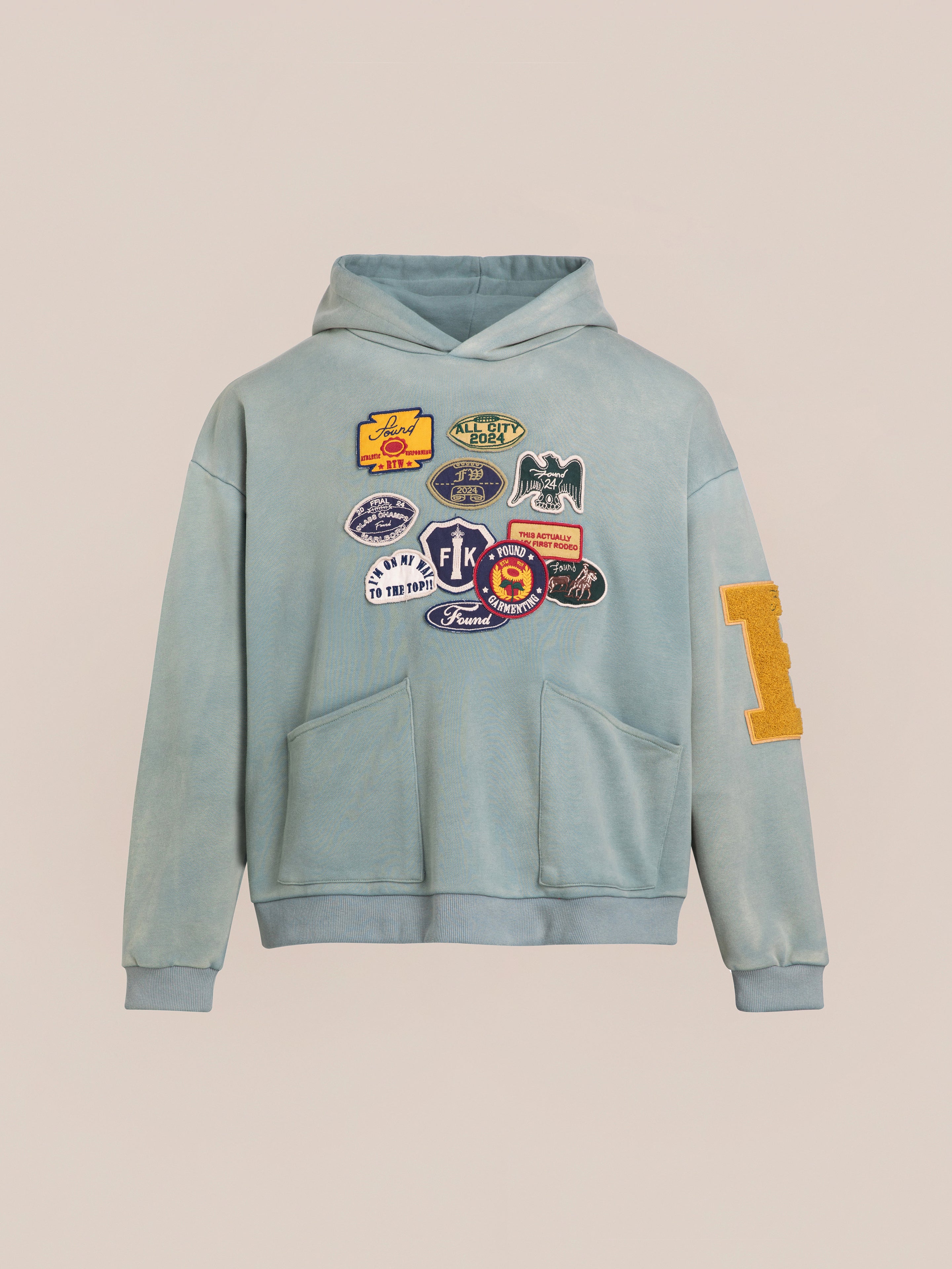 The Multi Patch Hoodie by FOUND, crafted from vintage-inspired French Terry cotton, features a light blue hue with a front pocket and strings. Various colorful patches and logos adorn the front, complemented by an "L" patch on the left sleeve.