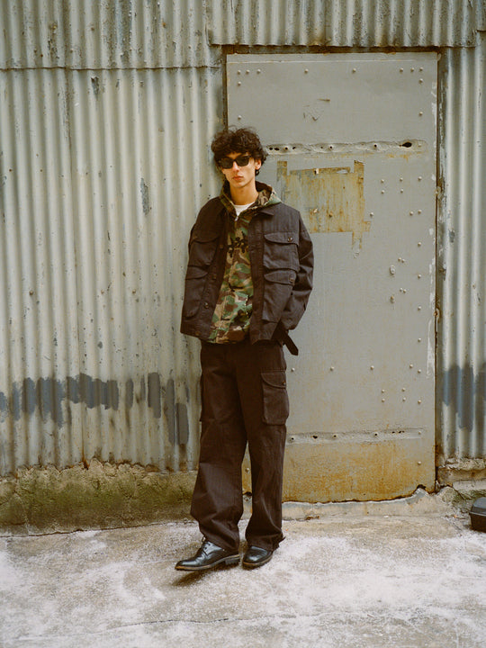 A person in sunglasses, wearing a black jacket and camo shirt, stands against a corrugated metal wall. They complete their look with FOUND's Utility Herringbone Cargo Pants, offering six roomy pockets for convenience and style.