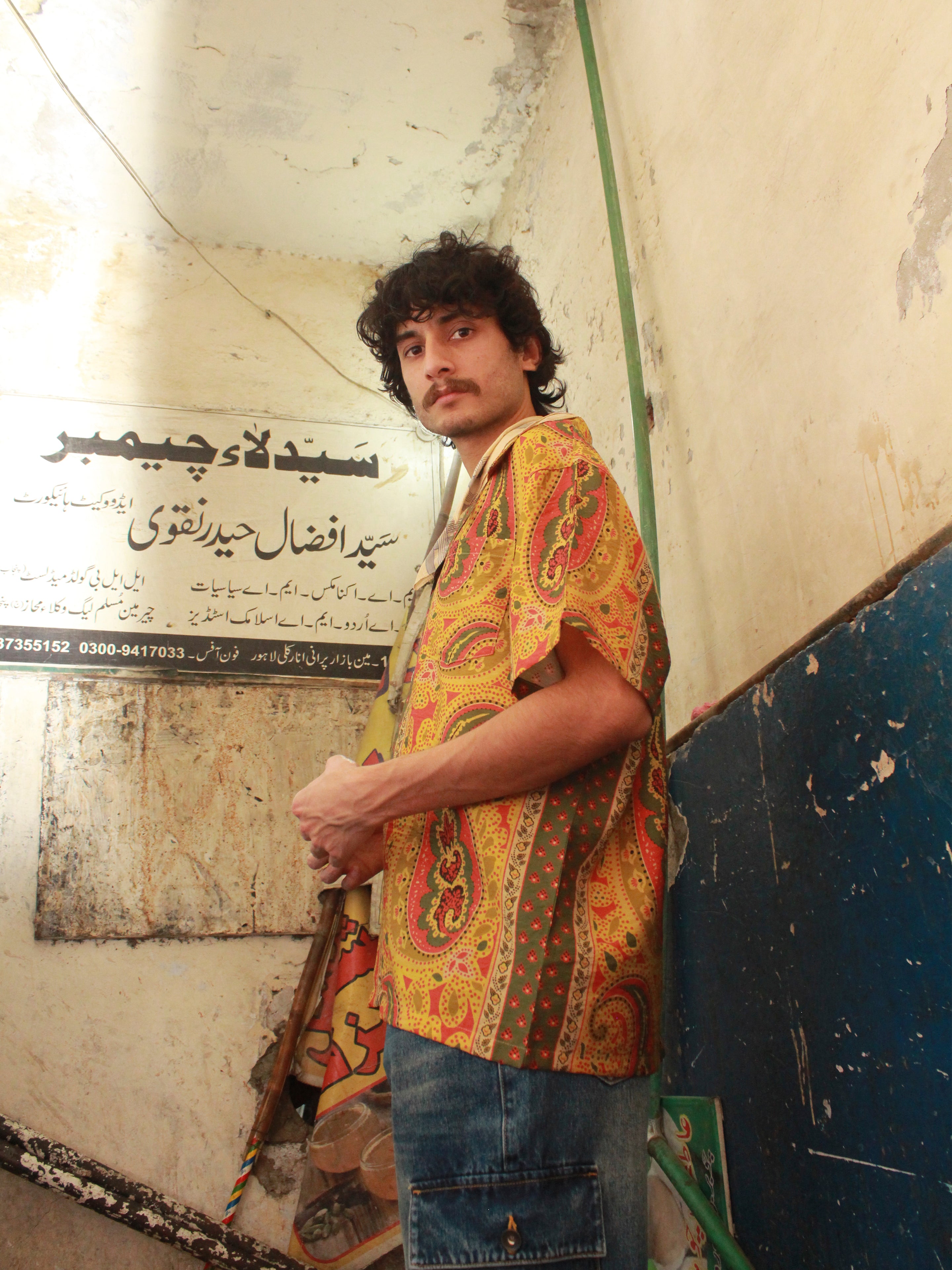 A man with curly hair and a mustache, dressed in a vibrant Pench Paisley SS Camp Shirt by Found, stands in a worn-out room with Arabic text on the wall behind him.