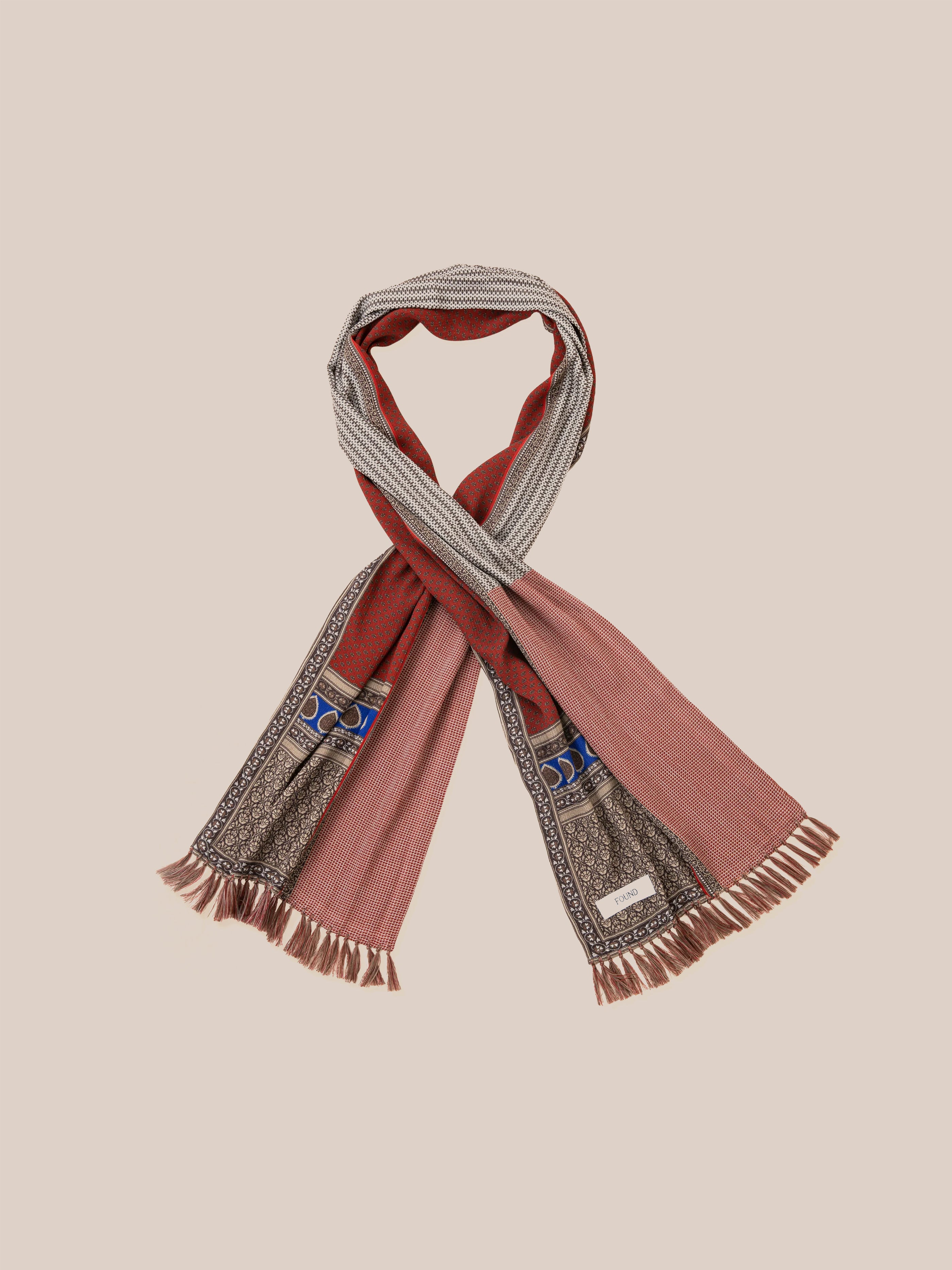 Introducing the Paisley Medley Scarf by Found—a chic fusion of Indo-Aryan designs in a vibrant mix of red, beige, and blue patterns. It includes hand-tied tassels and fringed edges, artfully set against a simple backdrop.