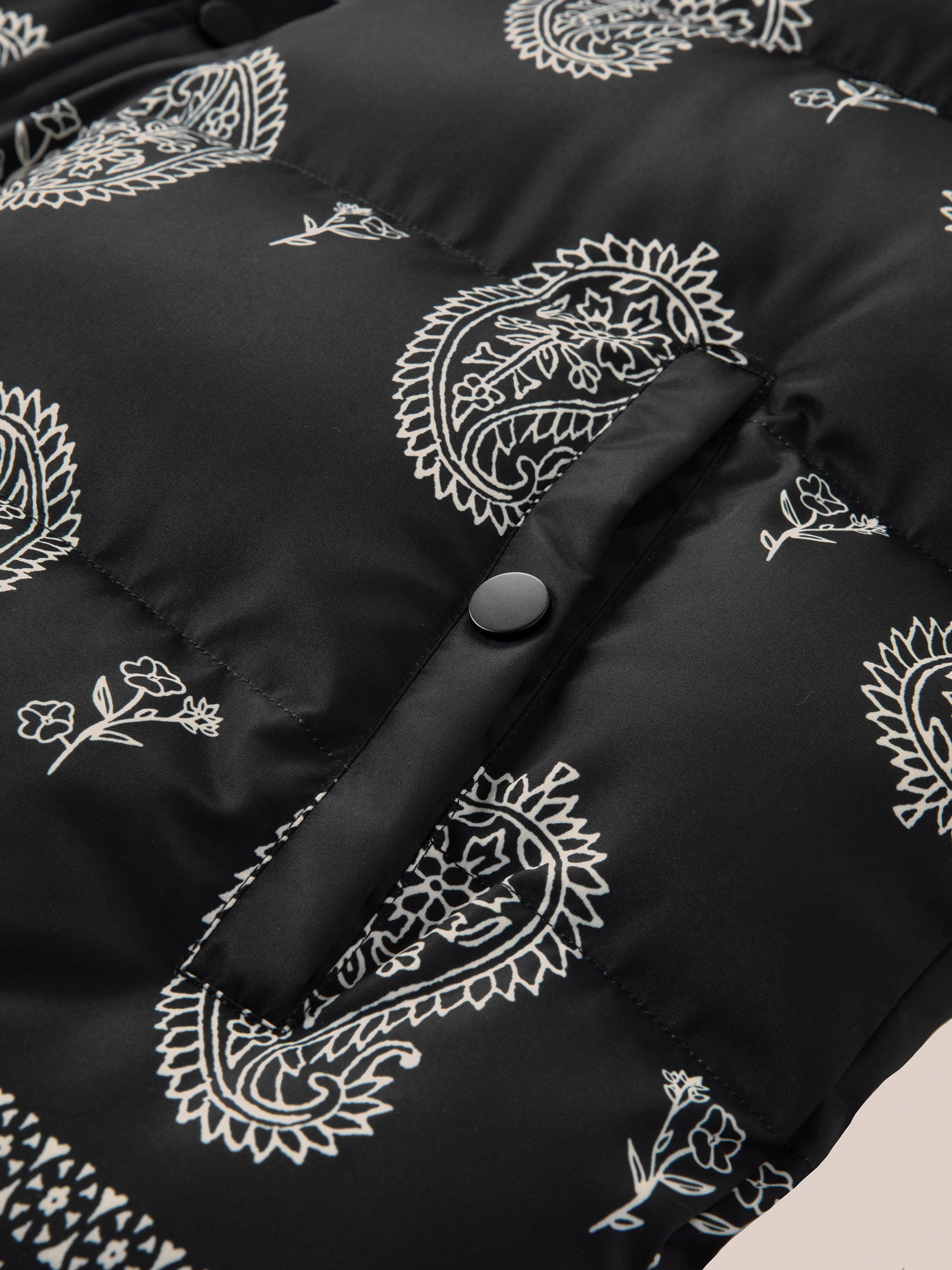 Close-up of the Paisley Puffer Jacket in Black by Profound, featuring white bandana paisley and floral patterns with a visible button on its padded nylon shell.