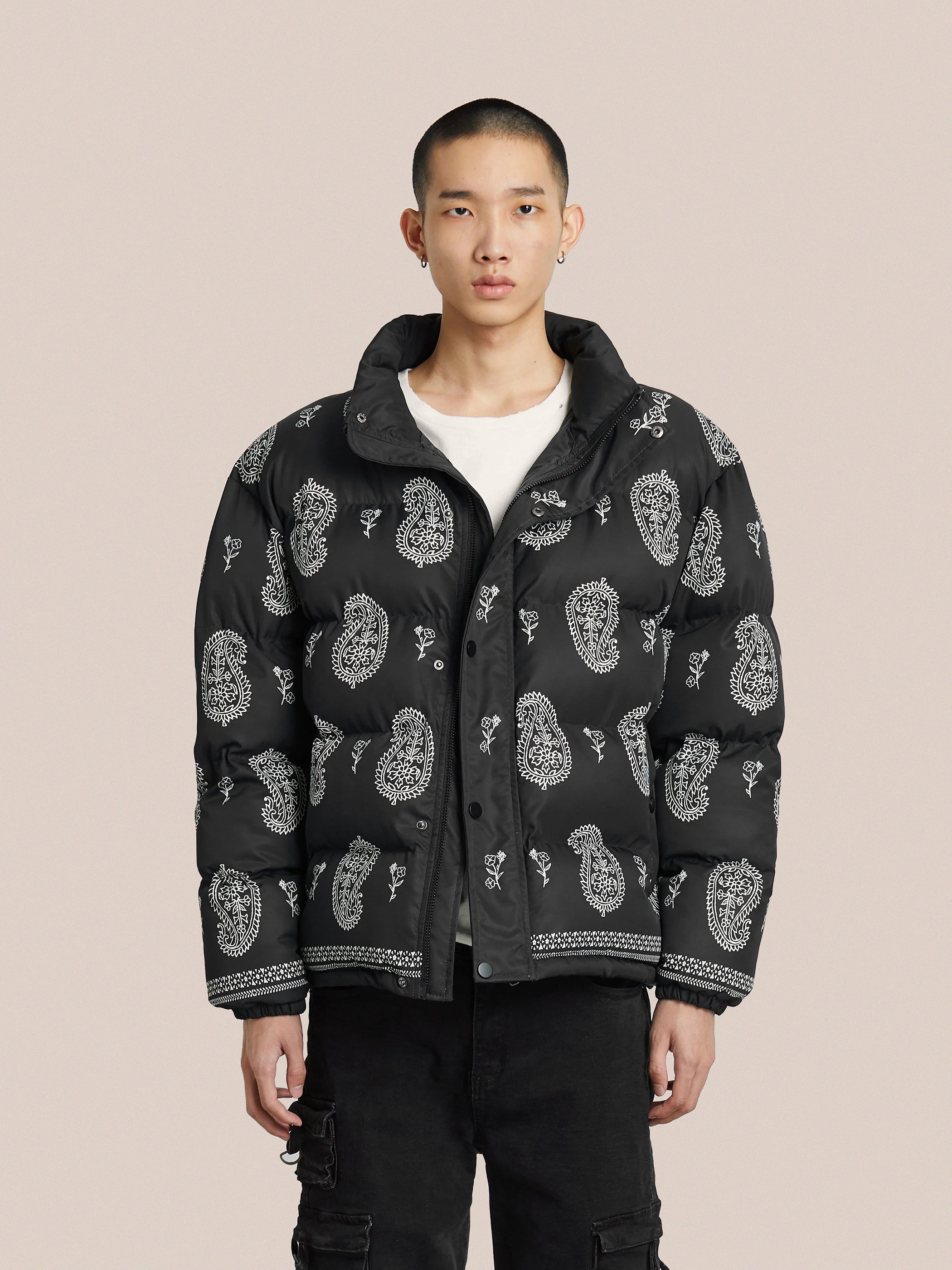 A person stands against a plain background wearing the Paisley Puffer Jacket in Black from Profound, featuring a striking black bandana paisley pattern on a nylon shell, paired with a crisp white shirt and sleek black pants.