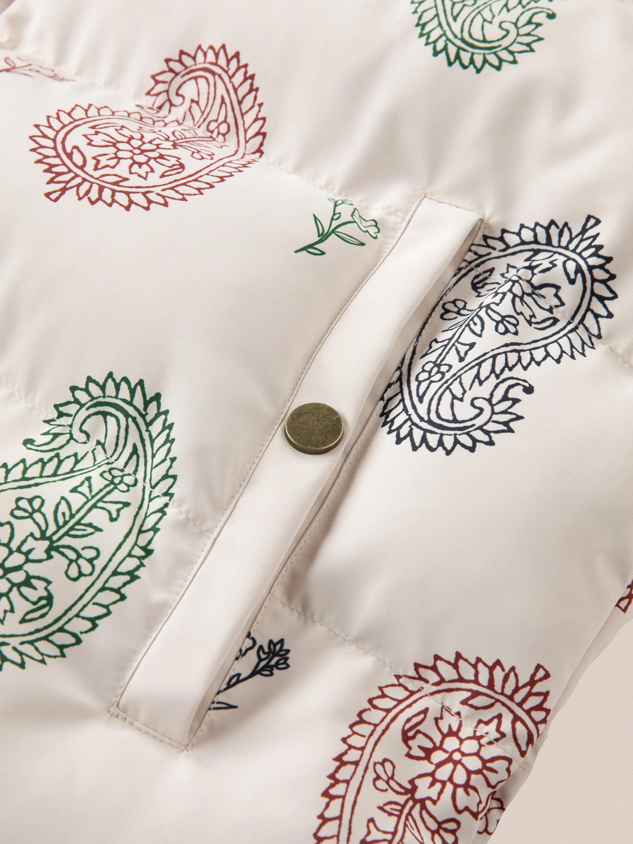 Close-up of a white fabric with a colorful paisley pattern in red, green, and black on the Paisley Puffer Jacket in Cream by FOUND, featuring a single brass snap button on a flap.