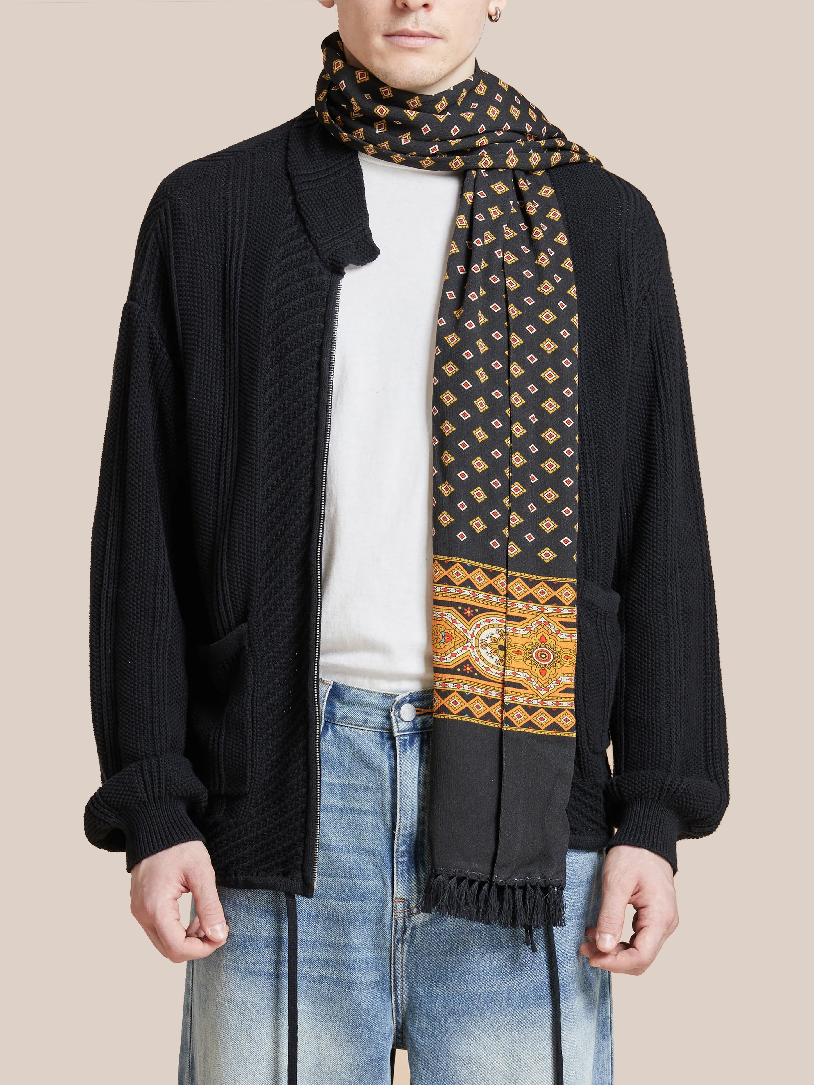 Person wearing a black cardigan, white shirt, and blue jeans is accessorized with the Found Pastures Print Scarf. This scarf features geometric and floral designs and is elegantly finished with hand-tied tassels for a charming touch.