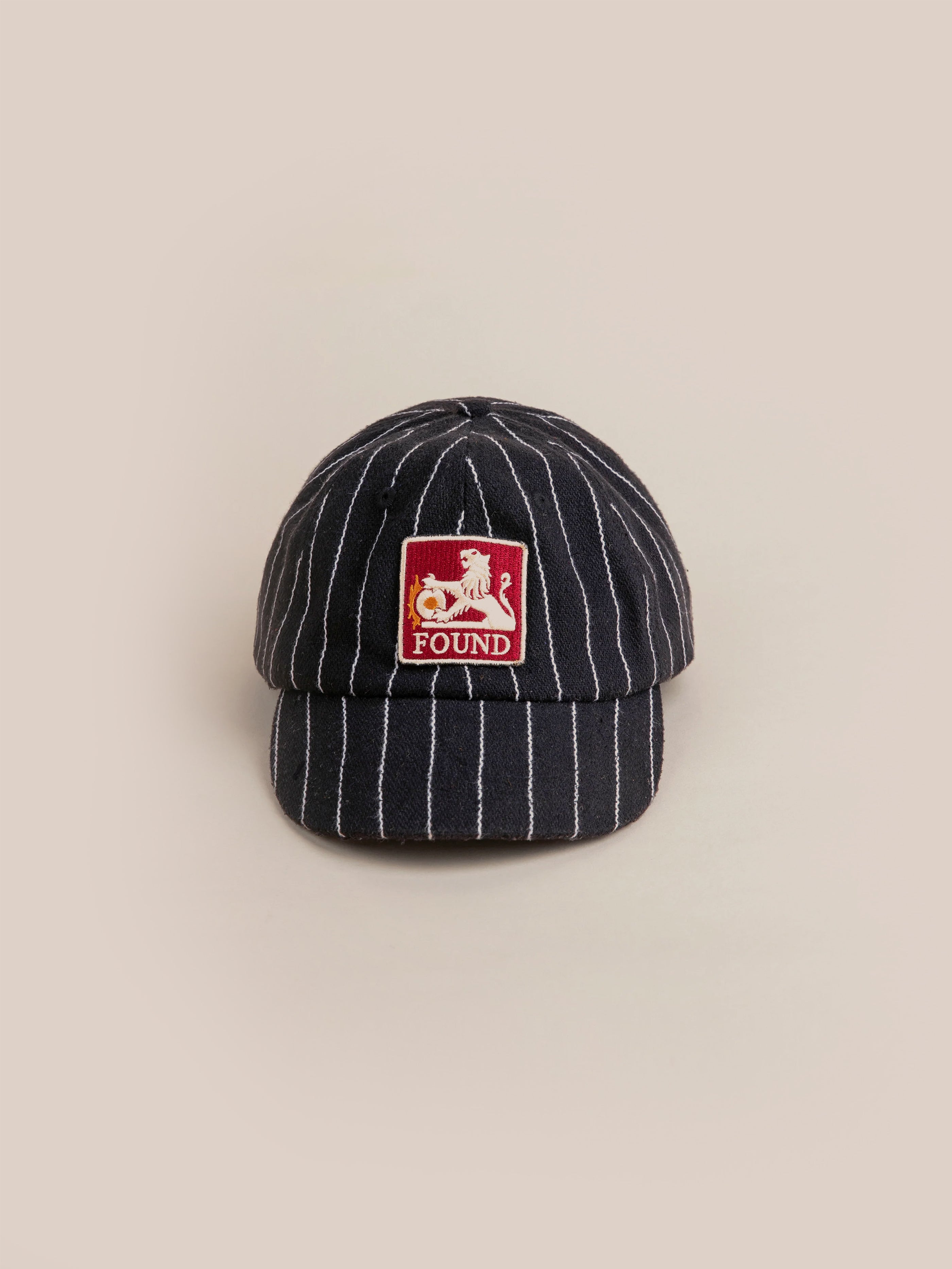 The Pinstripe Lion Cap by FOUND showcases a black and white pinstripe design, highlighted by a bold red square patch on the front that displays a cat and the word "FOUND." It also features an embroidered lion logo, adding a touch of distinctive charm.