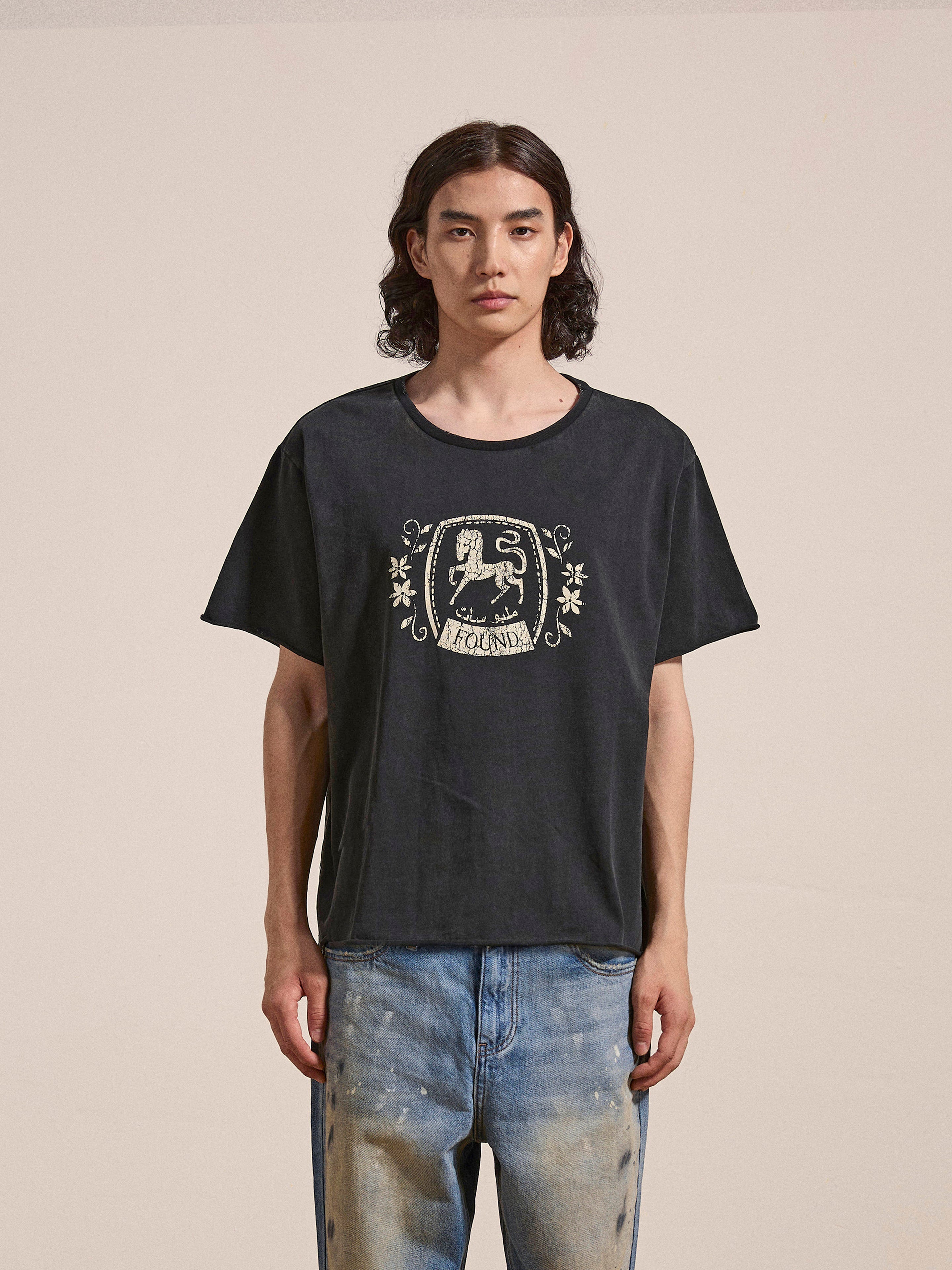 A person with long hair, wearing a FOUND Horse Logo Print Tee featuring a crest design, made from 100% cotton and treated with an enzyme wash, paired with light blue jeans, stands against a plain background.