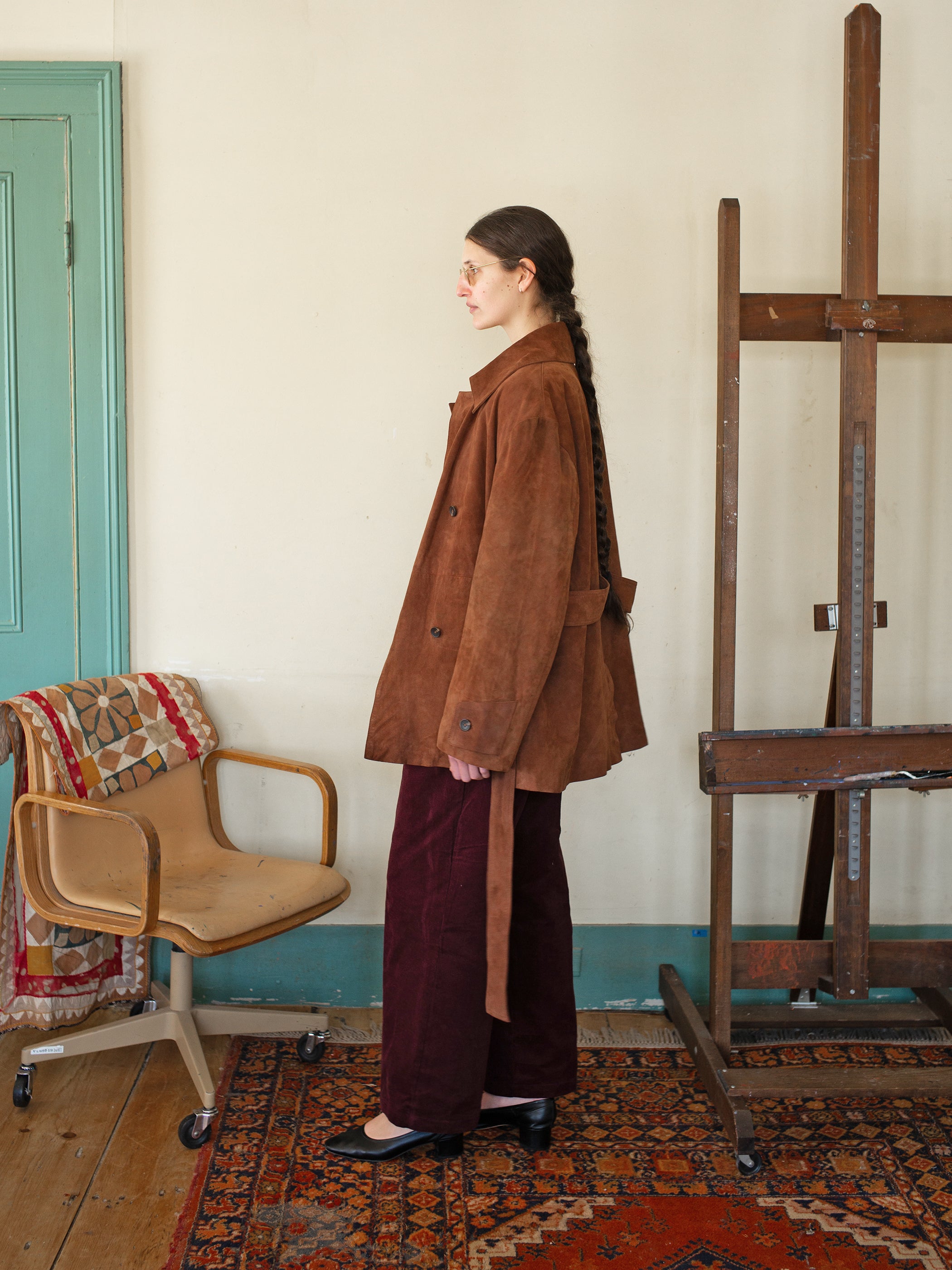 A person in FOUND's Double Breasted Suede Leather Jacket and red pants stands beside a wooden easel. A patterned fabric chair sits on a rug, enhancing the charm of the ethically produced space.