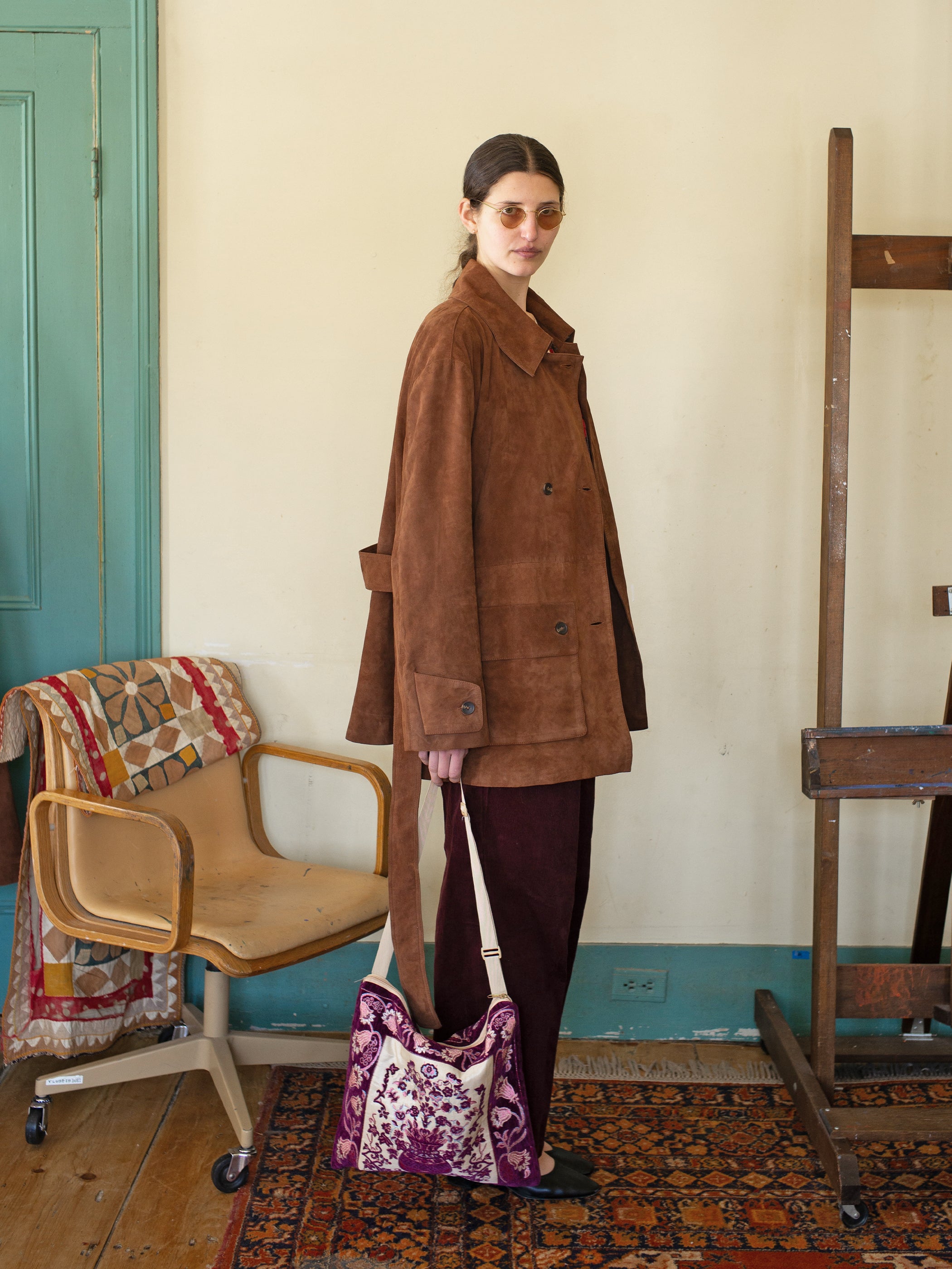 A person wearing the FOUND Double Breasted Suede Leather Jacket and sunglasses holds a floral bag, standing next to a chair draped with a patterned blanket in a room with light walls and a green door.