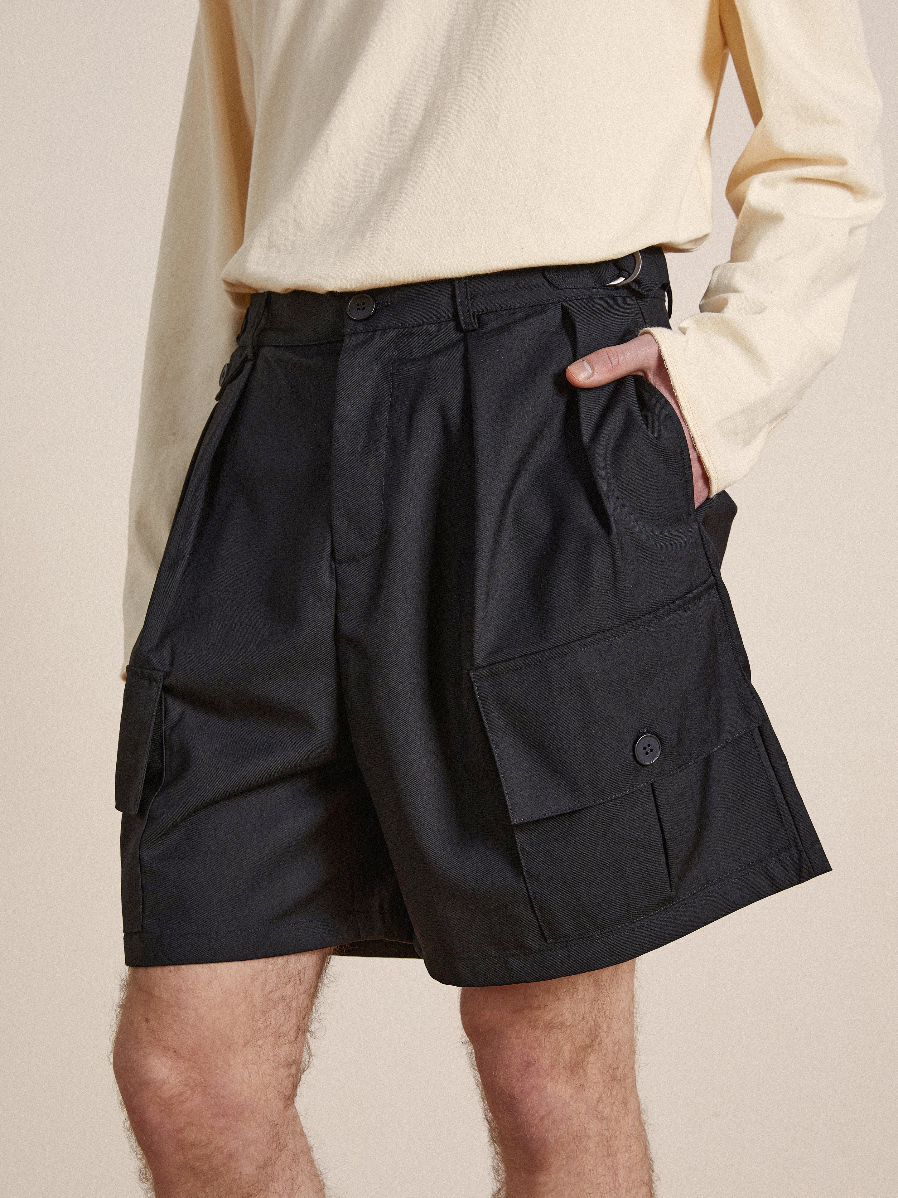 A person wearing a beige top and black Found Pleated Pocket Cargo Shorts stands with one hand in the pocket, focusing on the shorts' details and design.