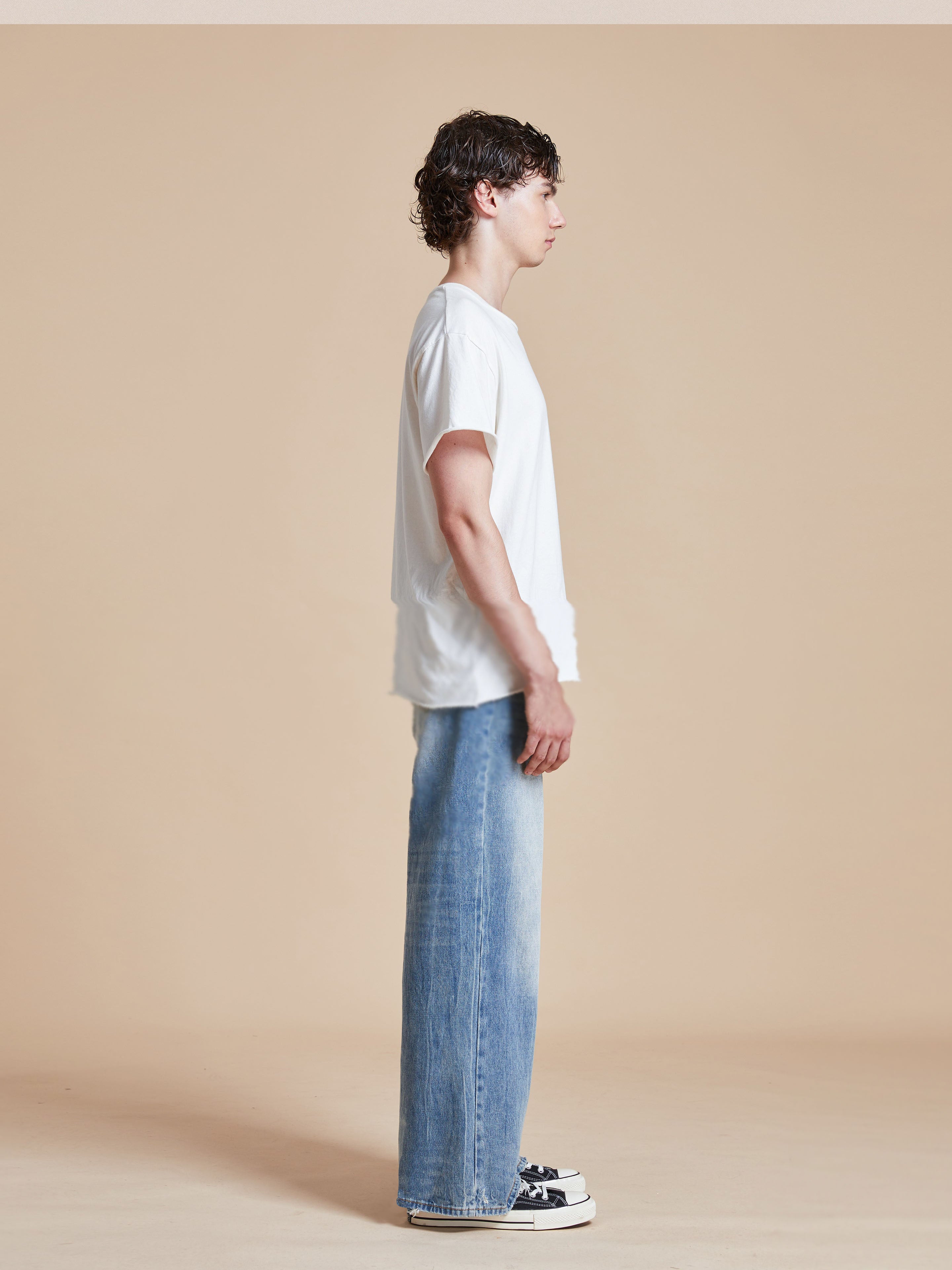 A person with curly hair stands sideways against a beige background, wearing a white t-shirt, Lacy Baggy Jeans by Found, and sneakers.