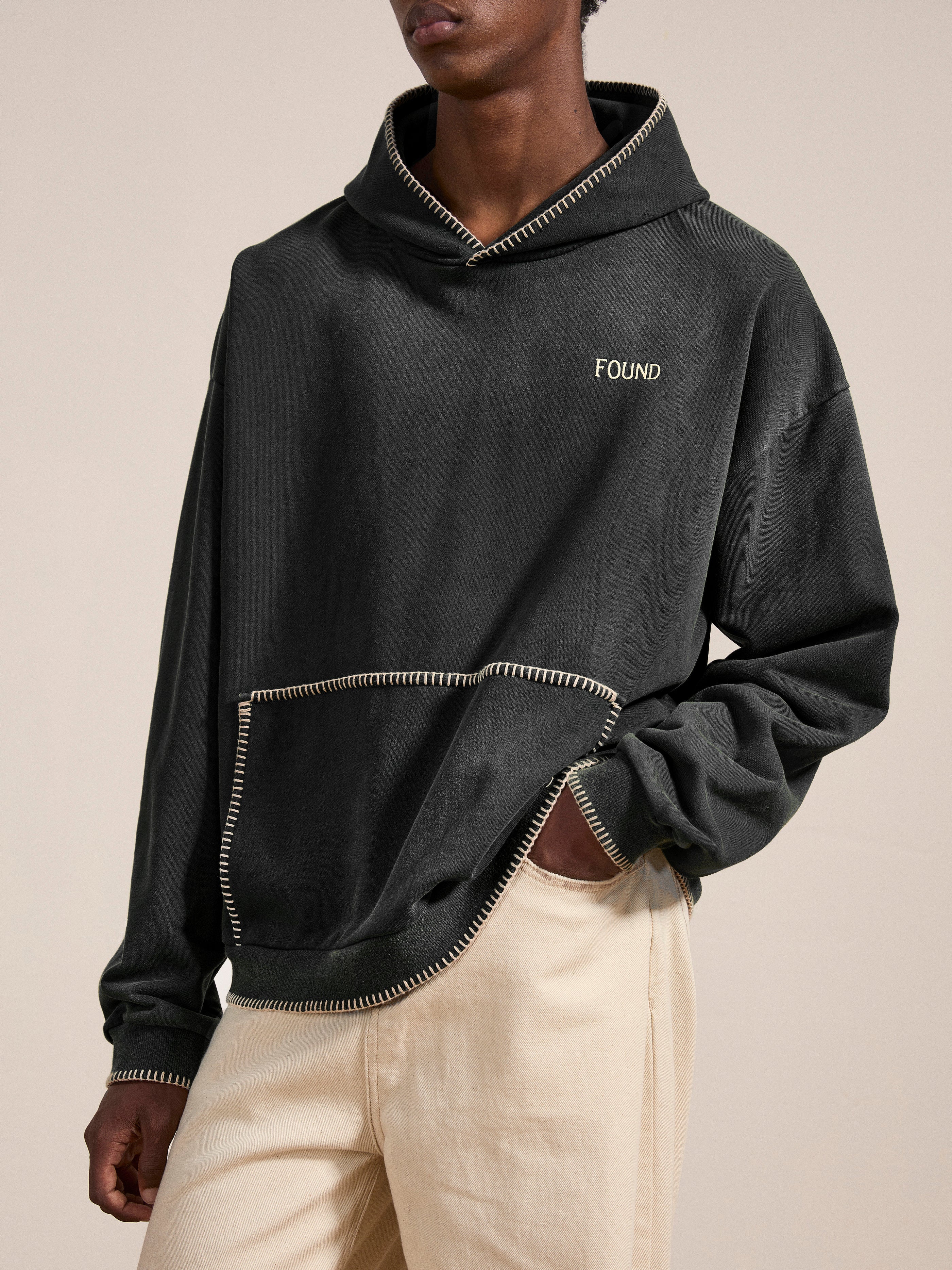 A person wearing an oversized, vintage black "Stitched Border Distressed Logo Hoodie" by FOUND and beige pants stands against a neutral background.