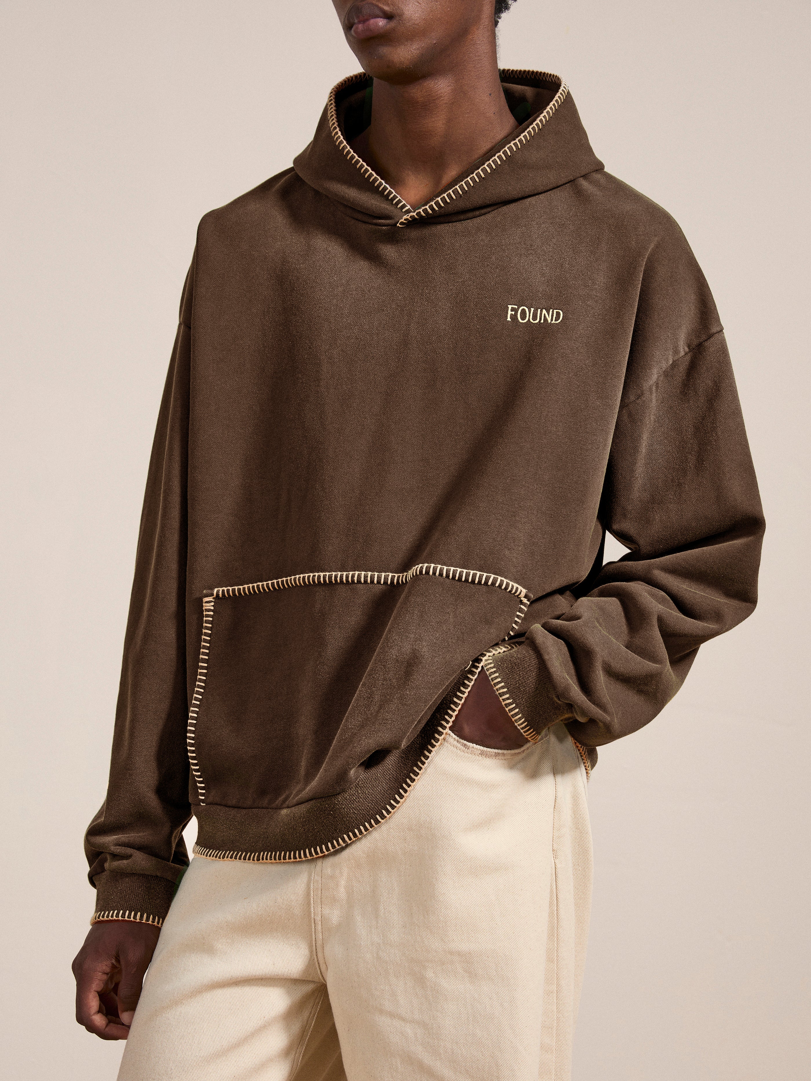 A person wearing a Stitched Border Distressed Logo Hoodie by FOUND, paired with beige pants, standing against a neutral background.