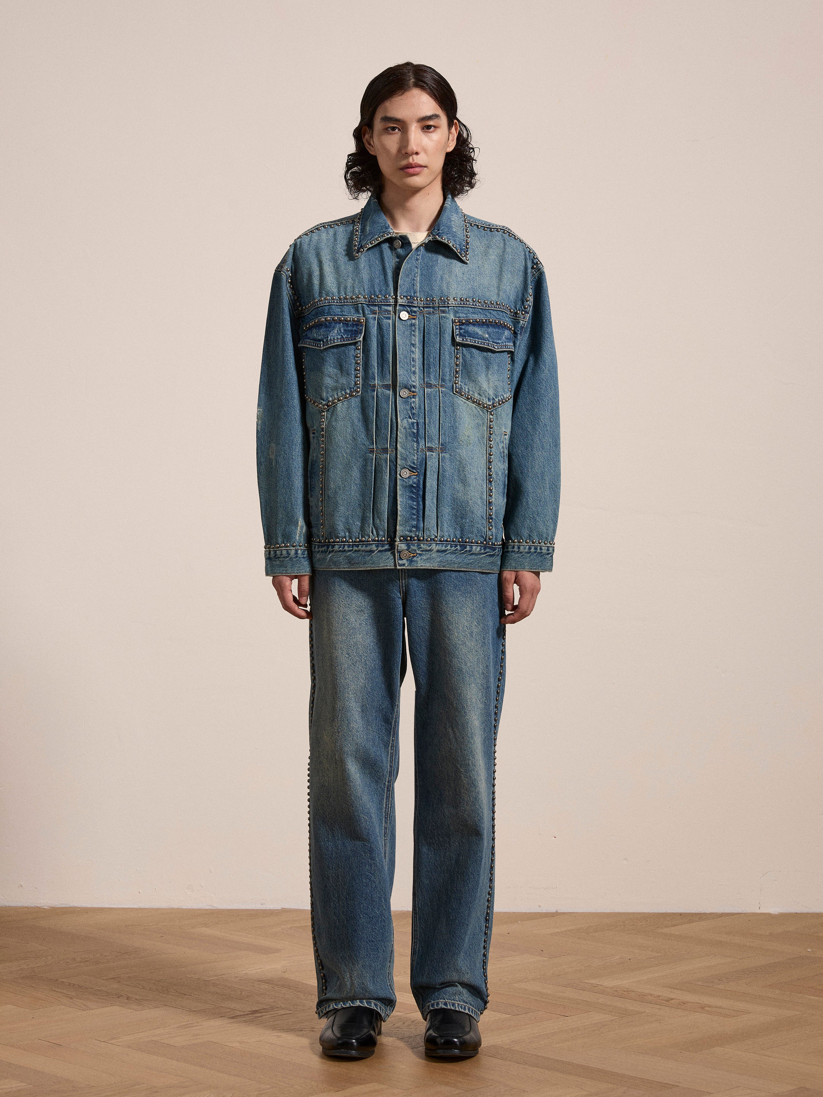 Person standing on a wooden floor wearing an oversized, boxy fit FOUND Azar Studded Denim Trucker Jacket and jeans in a vintage blue wash, black shoes, and having long dark hair. The background is a plain, light-colored wall.