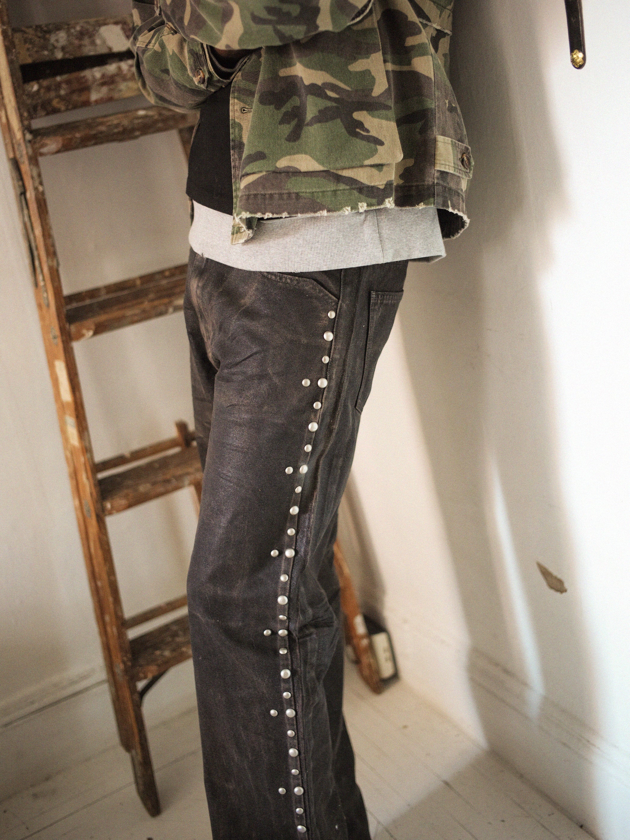 An individual wearing a camo jacket and FOUND Waxed Studded Jeans, inspired by vintage craftsmanship, stands near an indoor wooden ladder.