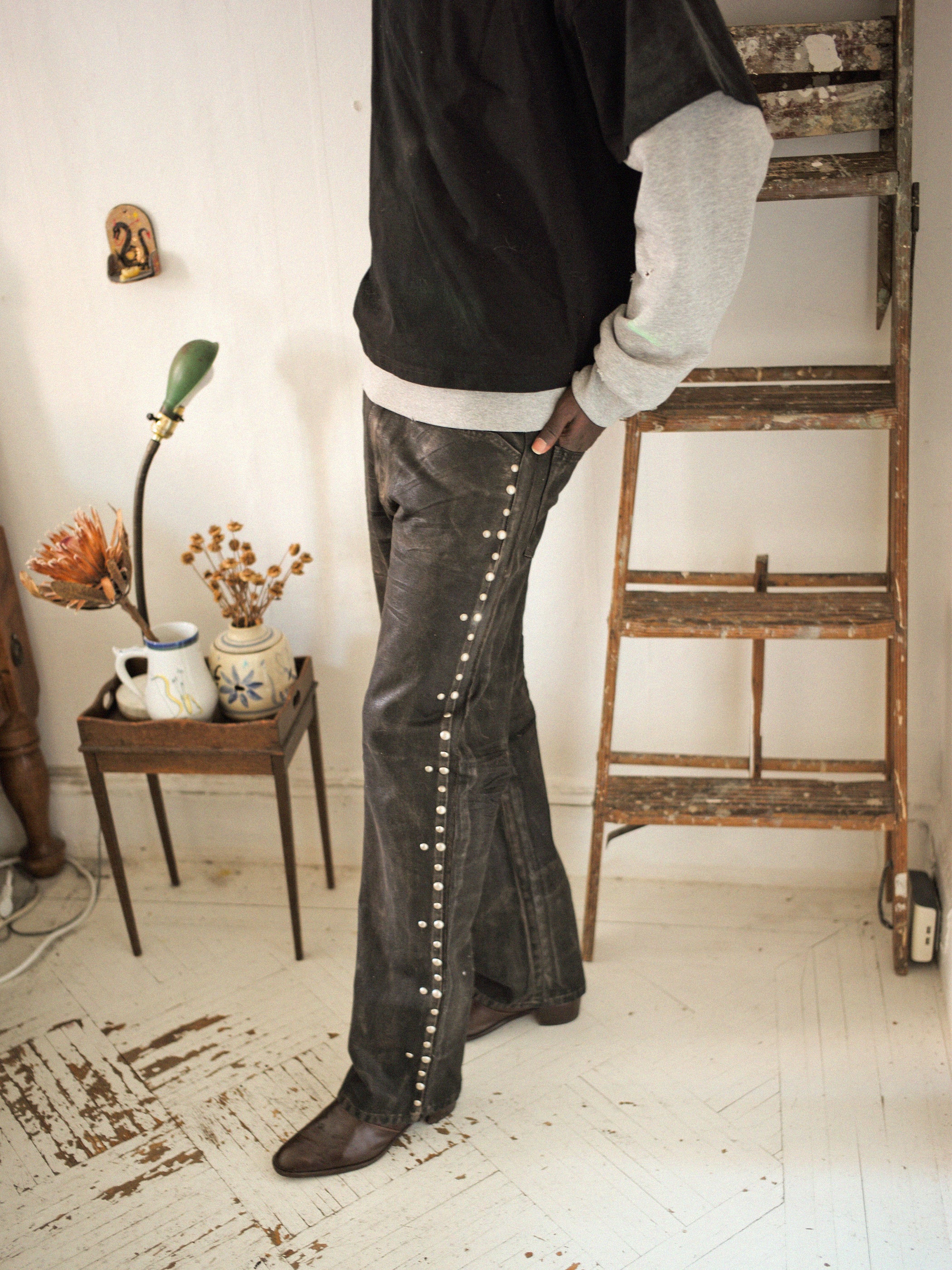 A person wearing FOUND's Waxed Studded Jeans in a black and gray outfit stands by an indoor wooden ladder. A table nearby displays a lamp, flowers, and mugs, with the floor reflecting vintage craftsmanship.