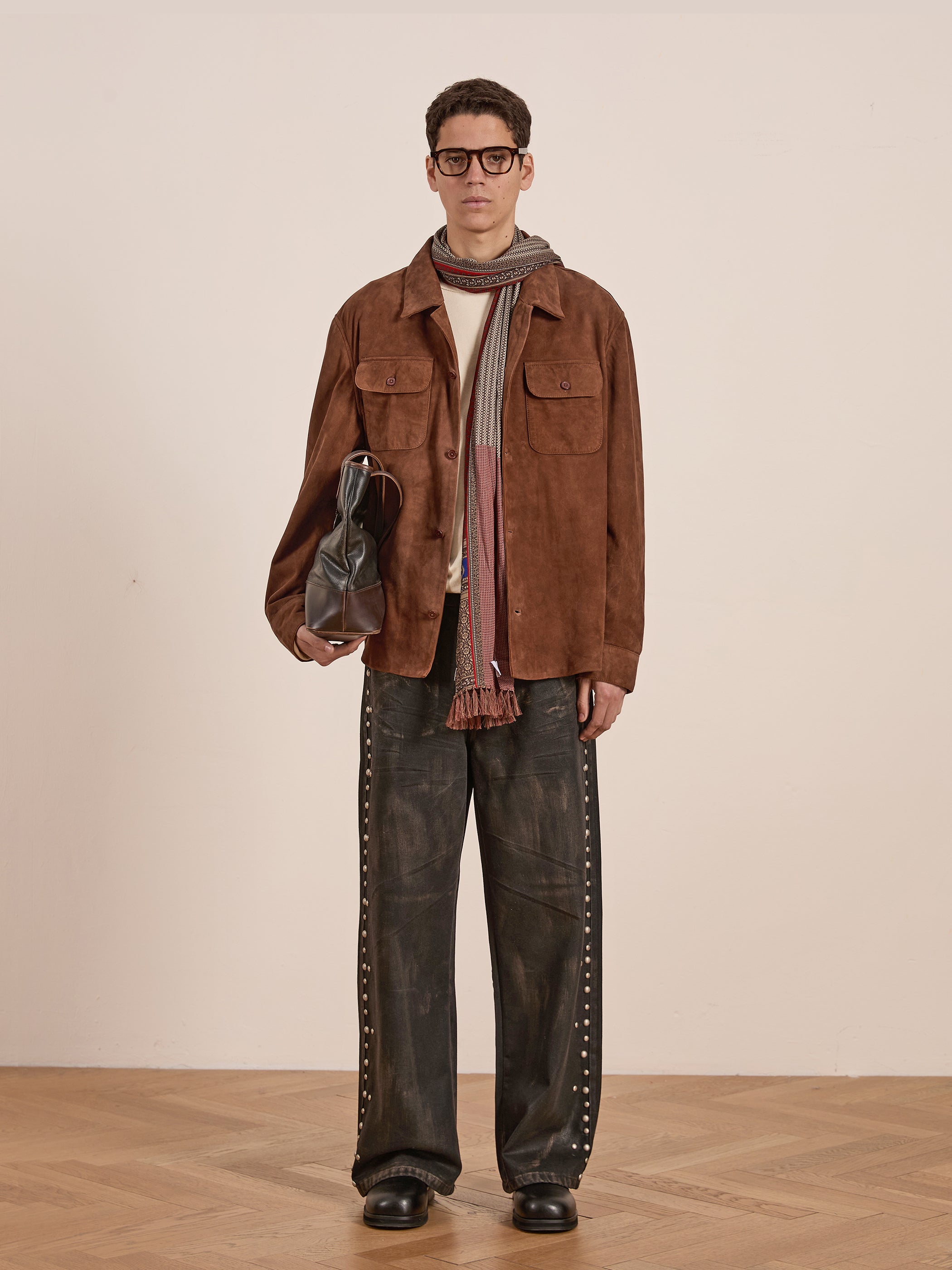 A person stands against a plain backdrop wearing a FOUND Suede Camp Shirt, patterned scarf, loose dark pants, and holding a black bag.