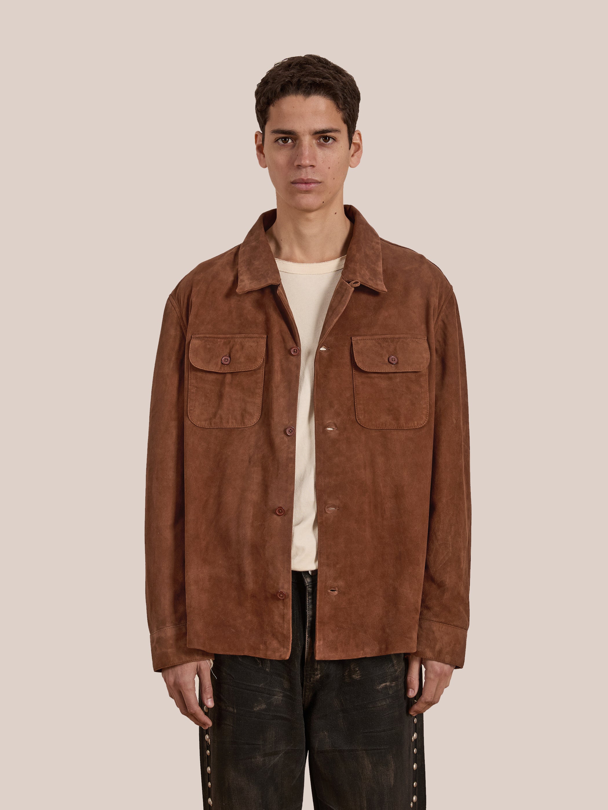 A person effortlessly rocks a rugged workwear style, wearing the Suede Camp Shirt from FOUND, crafted from genuine brown goat suede leather over a beige shirt, set against a plain background.