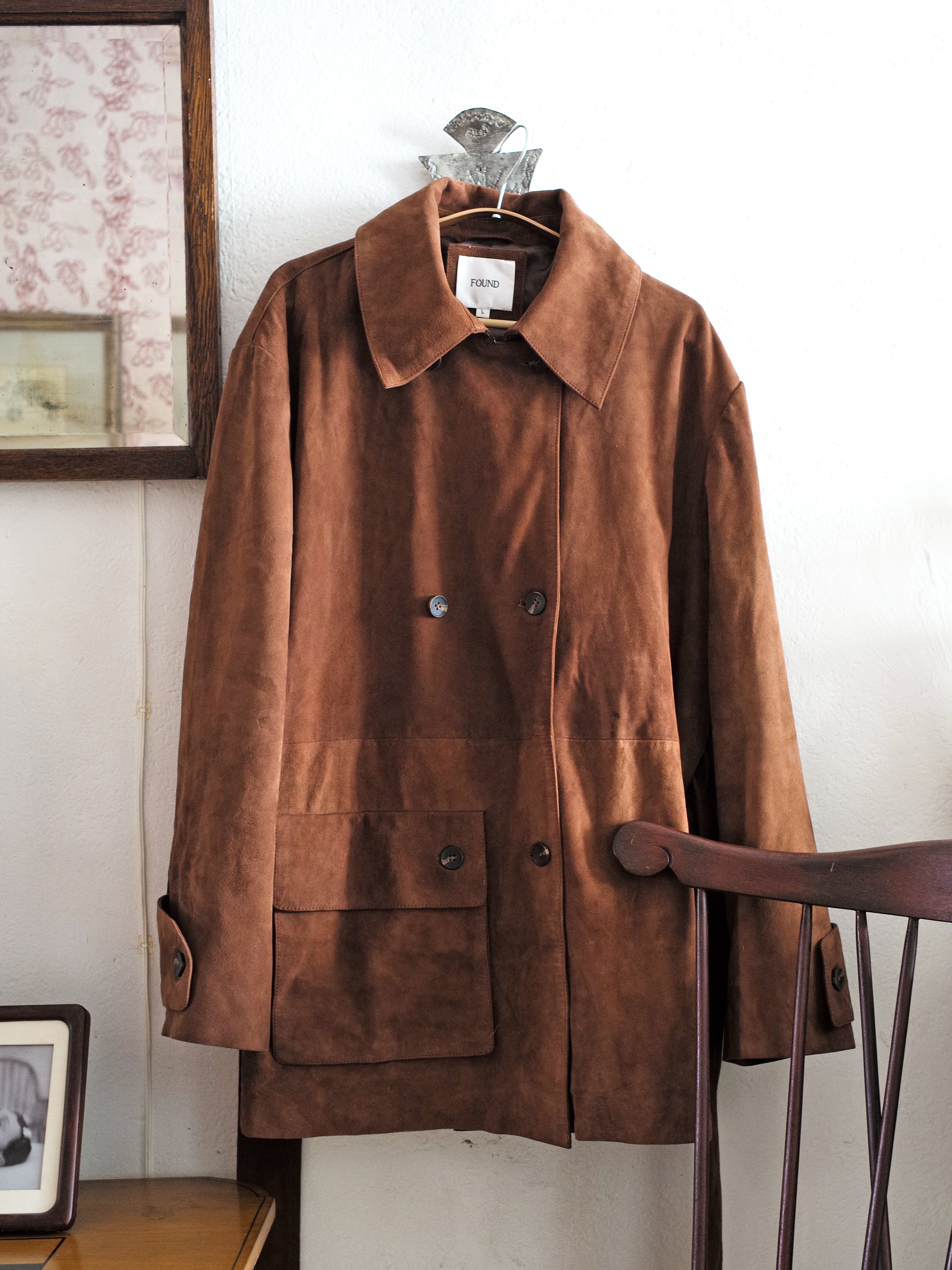 A Double Breasted Suede Leather Jacket by FOUND hangs on the wall beside a wooden chair, with framed pictures in the background.