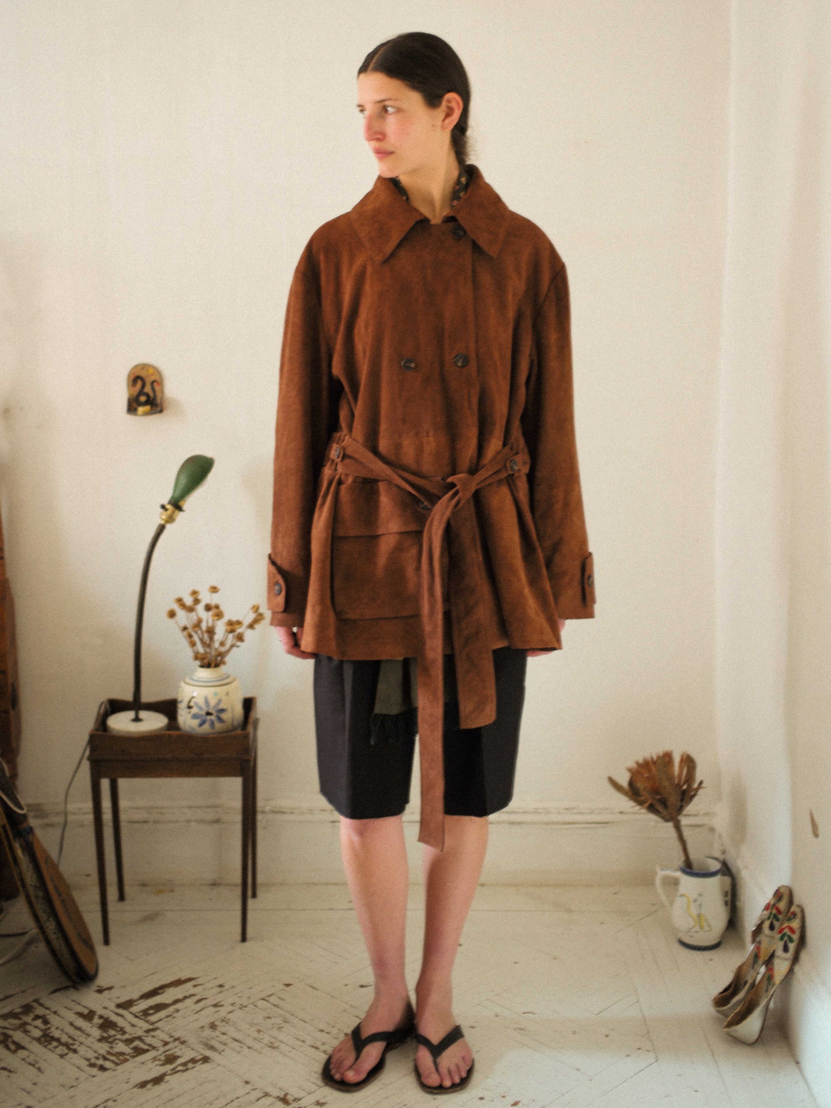 A person wearing a FOUND Double Breasted Suede Leather Jacket and black shorts stands in a room with a small table, lamp, and decorative items.