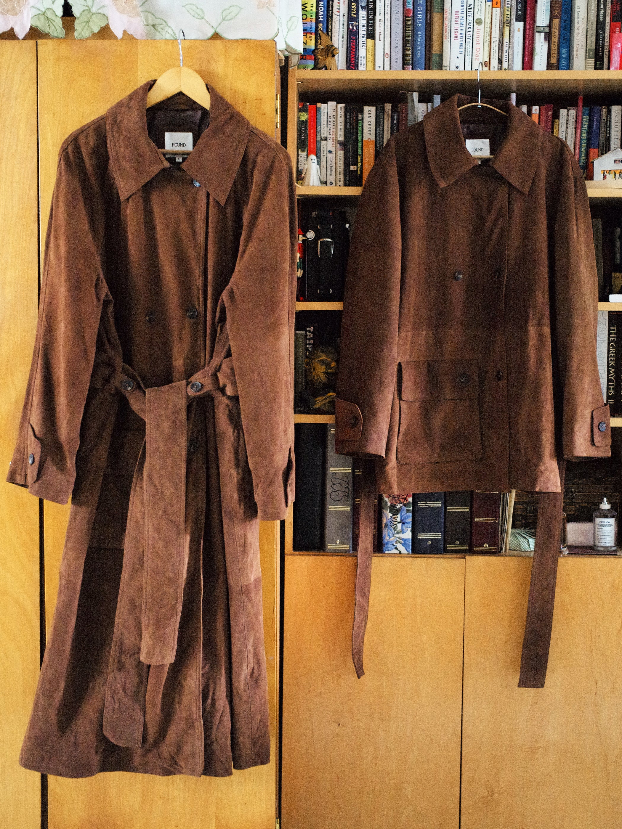 Two unisex brown suede leather coats hang in front of a bookshelf: one is a long trench coat from FOUND, the other a shorter jacket. Both are ethically crafted in Pakistan, combining style with conscious craftsmanship.