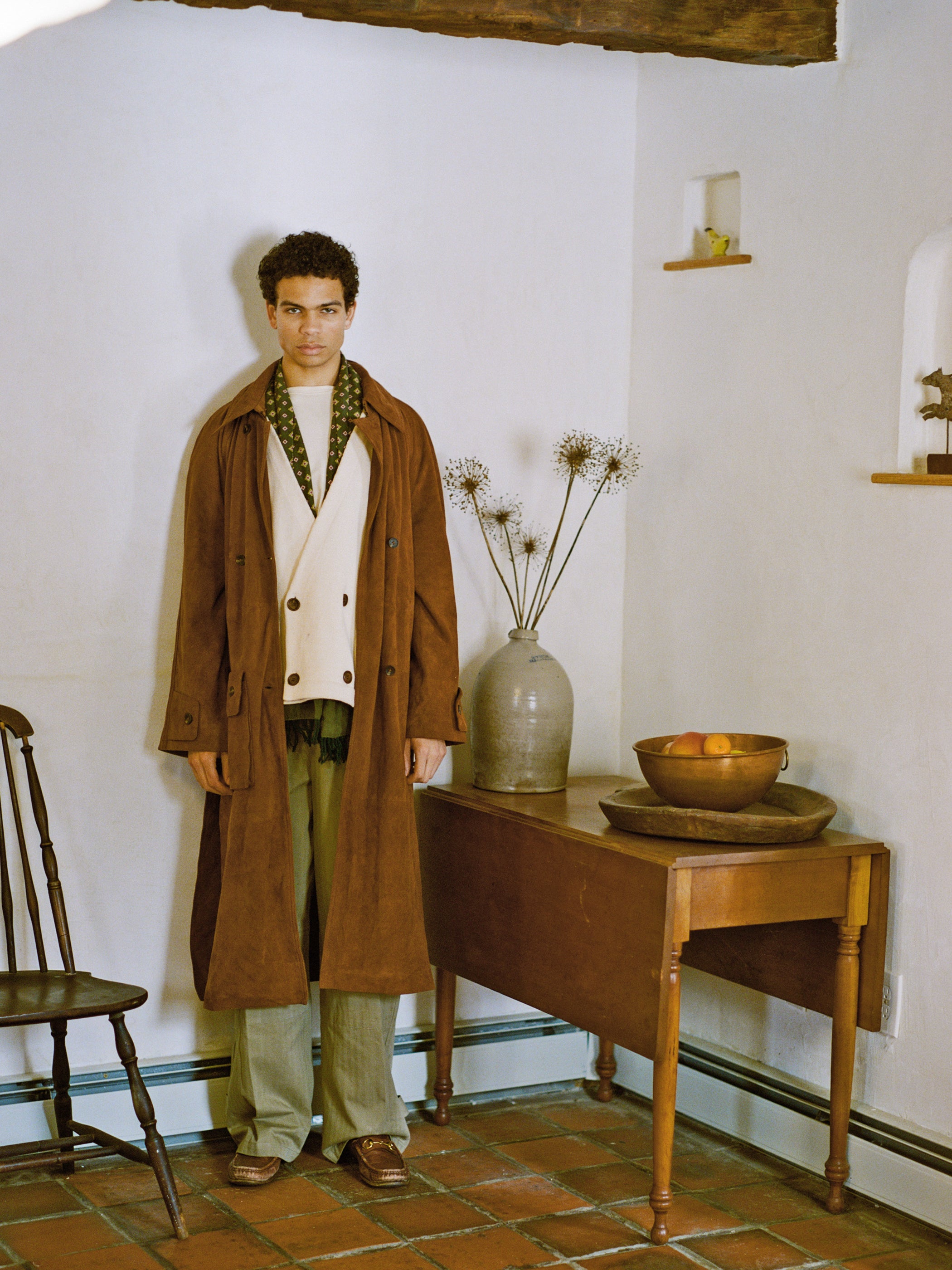 A person wearing a FOUND Suede Leather Trench Coat and green pants stands by a wooden table with a vase and fruit bowl, adding elegance to the room. This unisex piece is ethically crafted in Pakistan.