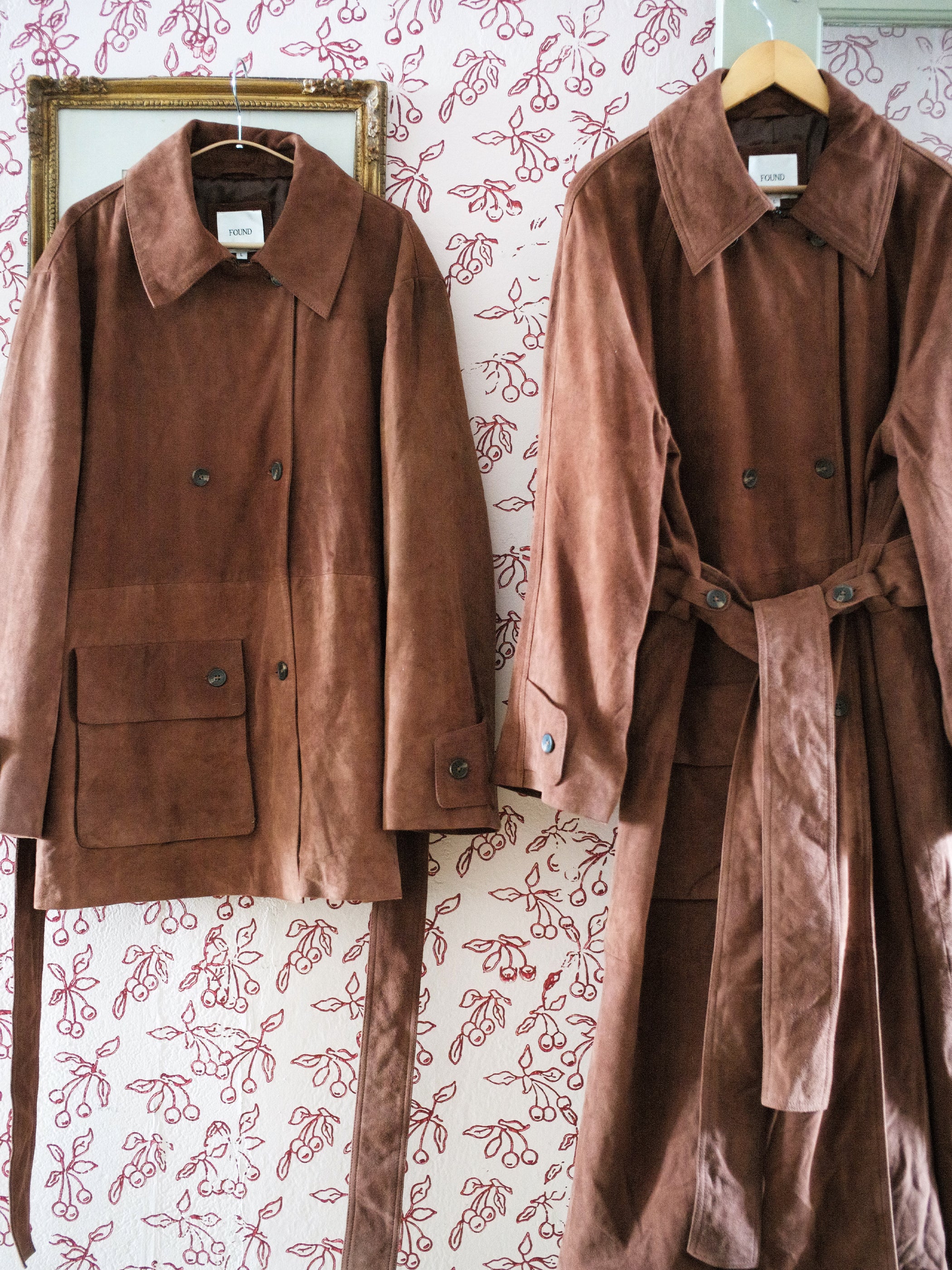 Two brown suede leather jackets hang against a white wall with red floral patterns. The jacket on the left is shorter with buttoned pockets, while the one on the right, from FOUND, is longer and double-breasted, featuring a stylish belt.