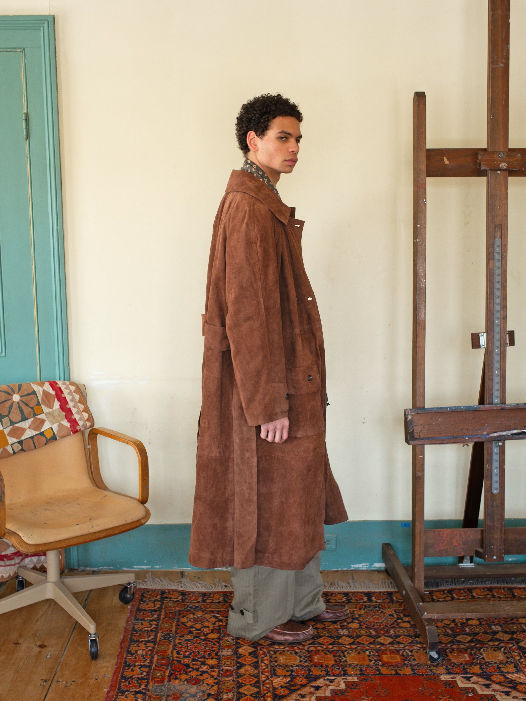 A person wearing a brown Suede Leather Trench Coat by FOUND, ethically crafted in Pakistan, stands sideways in a room with an easel, chair, and patterned rug.