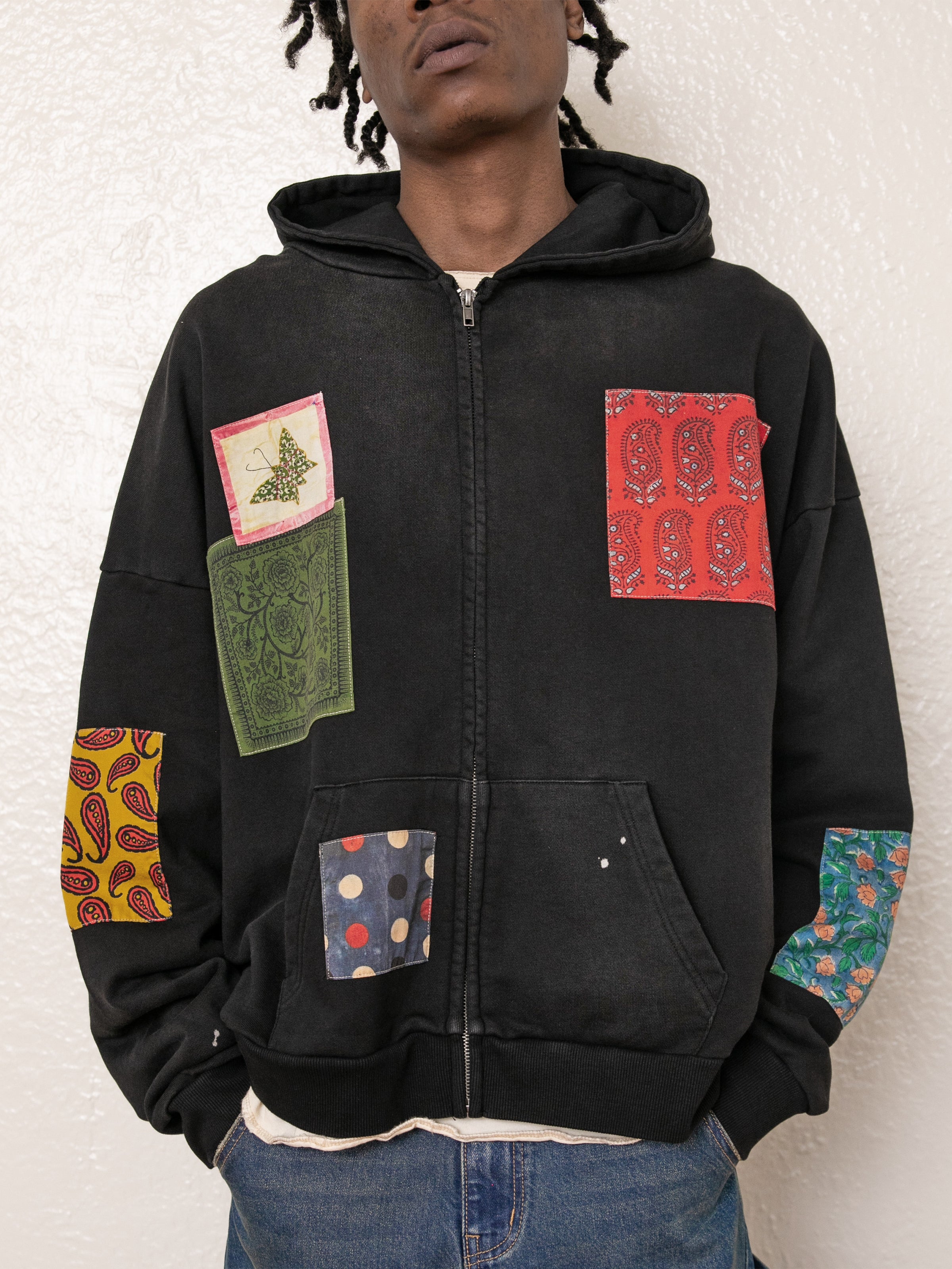 A person wearing the Tapestry Patchwork Zip Up Hoodie by FOUND, a vintage-inspired black hoodie adorned with vibrant patch designs, stands against a white wall.