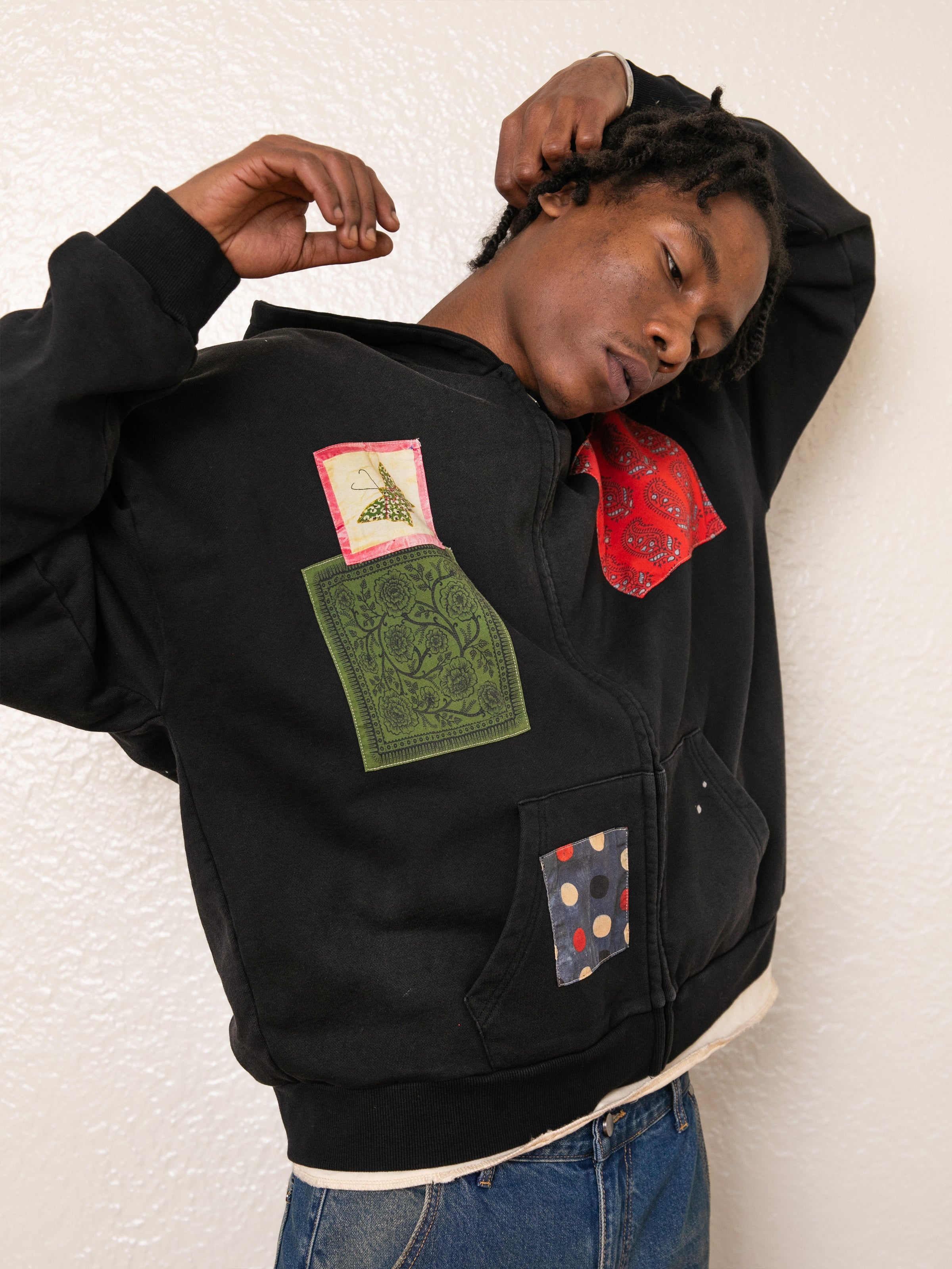 A person in a FOUND Tapestry Patchwork Zip Up Hoodie stands against an off-white wall, arms bent.