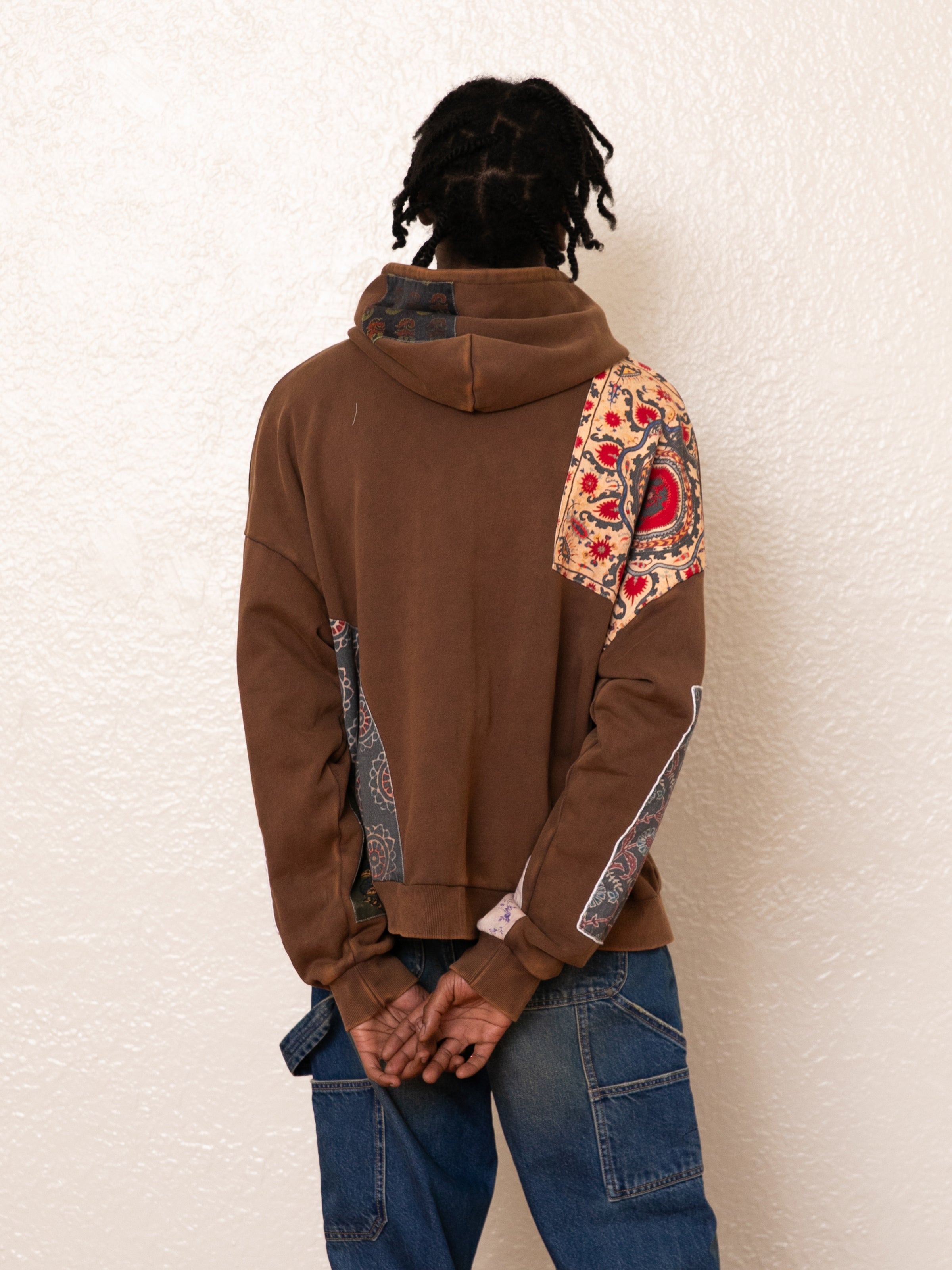 A person in an oversized beige FOUND Tapestry Patchwork Zip Up Hoodie and blue jeans stands facing away against a white textured wall.