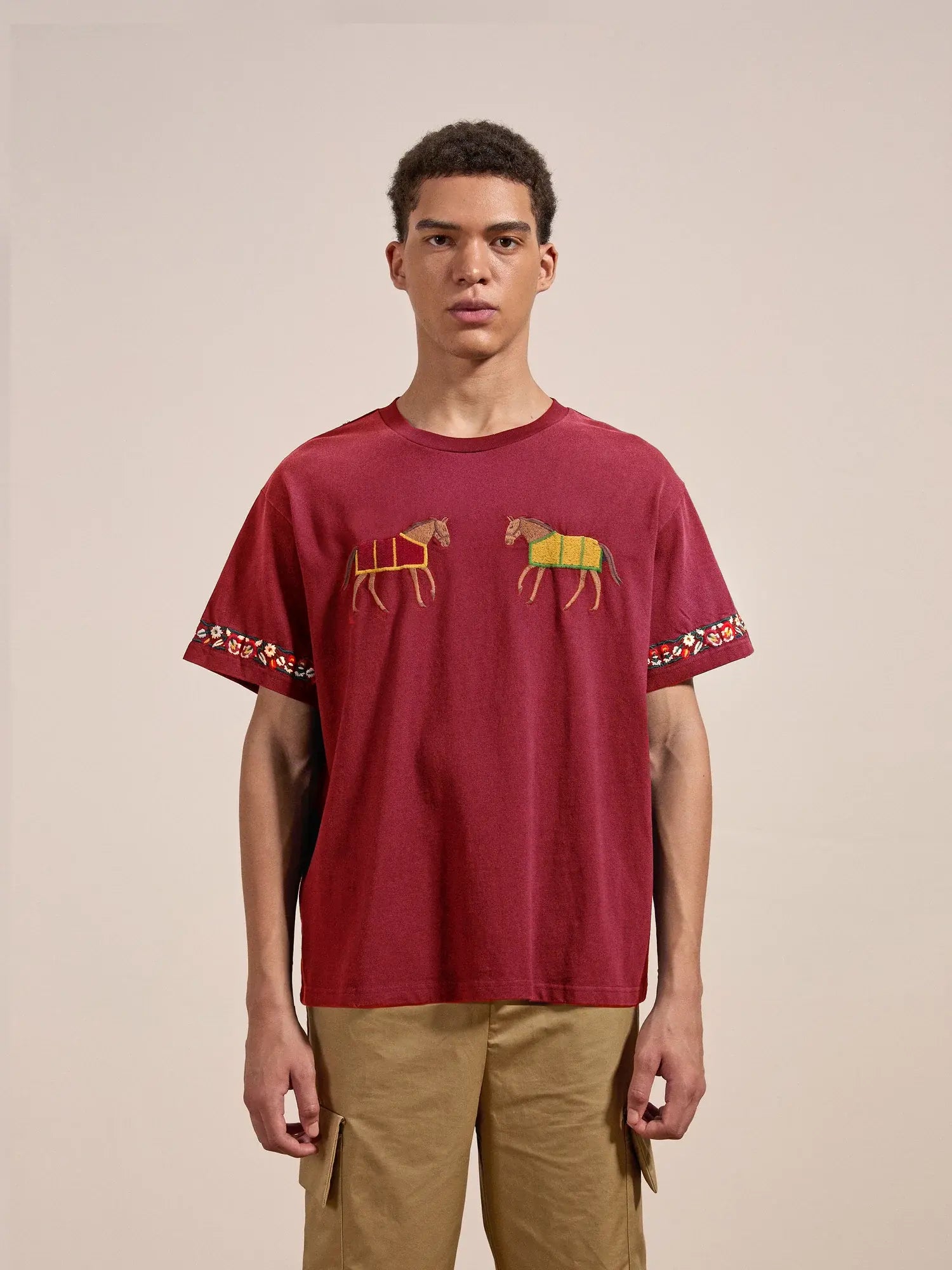 A person wearing the Horse Equine Tee by FOUND, which features colorful animal designs and detailed embroidery reminiscent of 70's jockey uniforms on the front, paired with khaki pants.